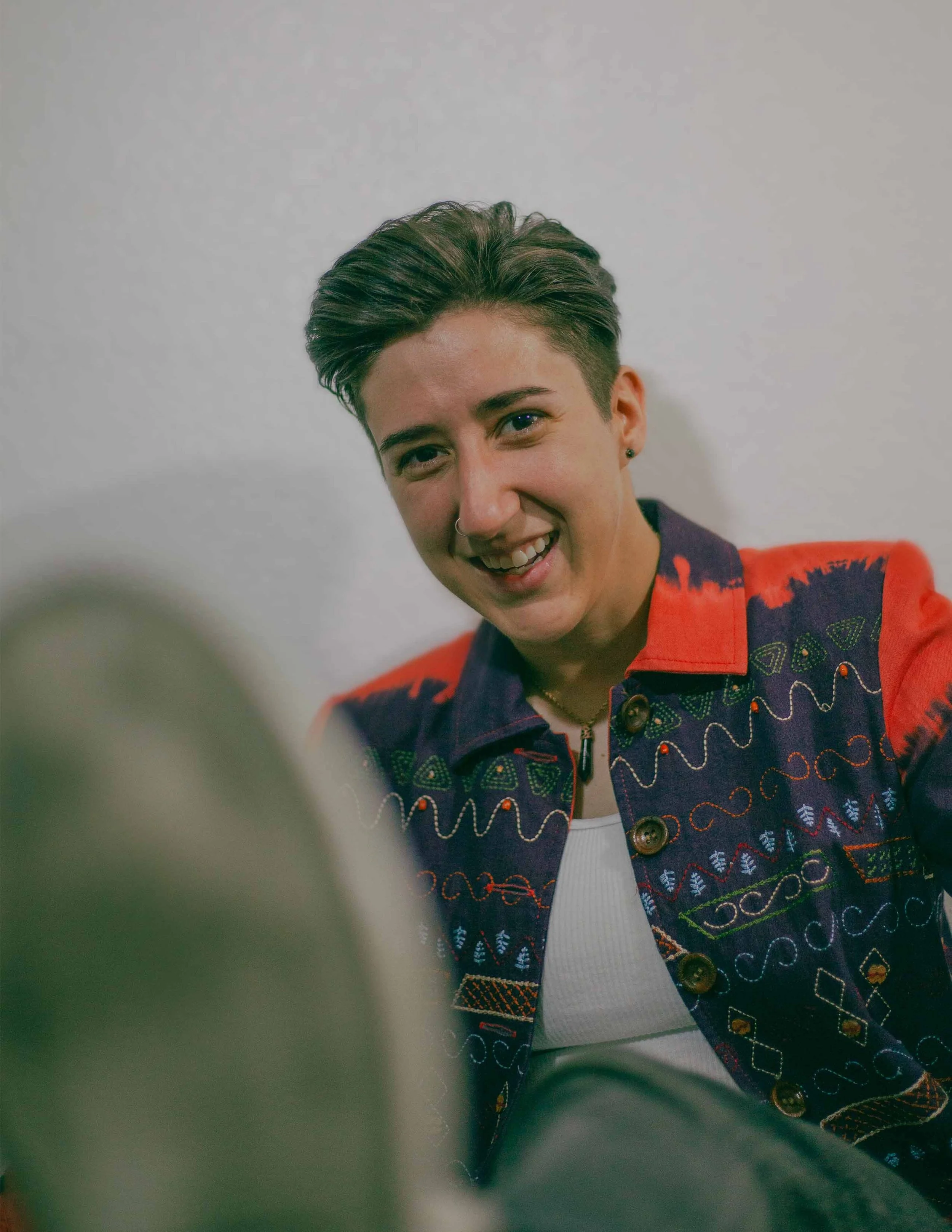 A woman with short, dark hair, smiling, wearing a multicolored embroidered jacket over a white shirt, sitting against a plain wall.