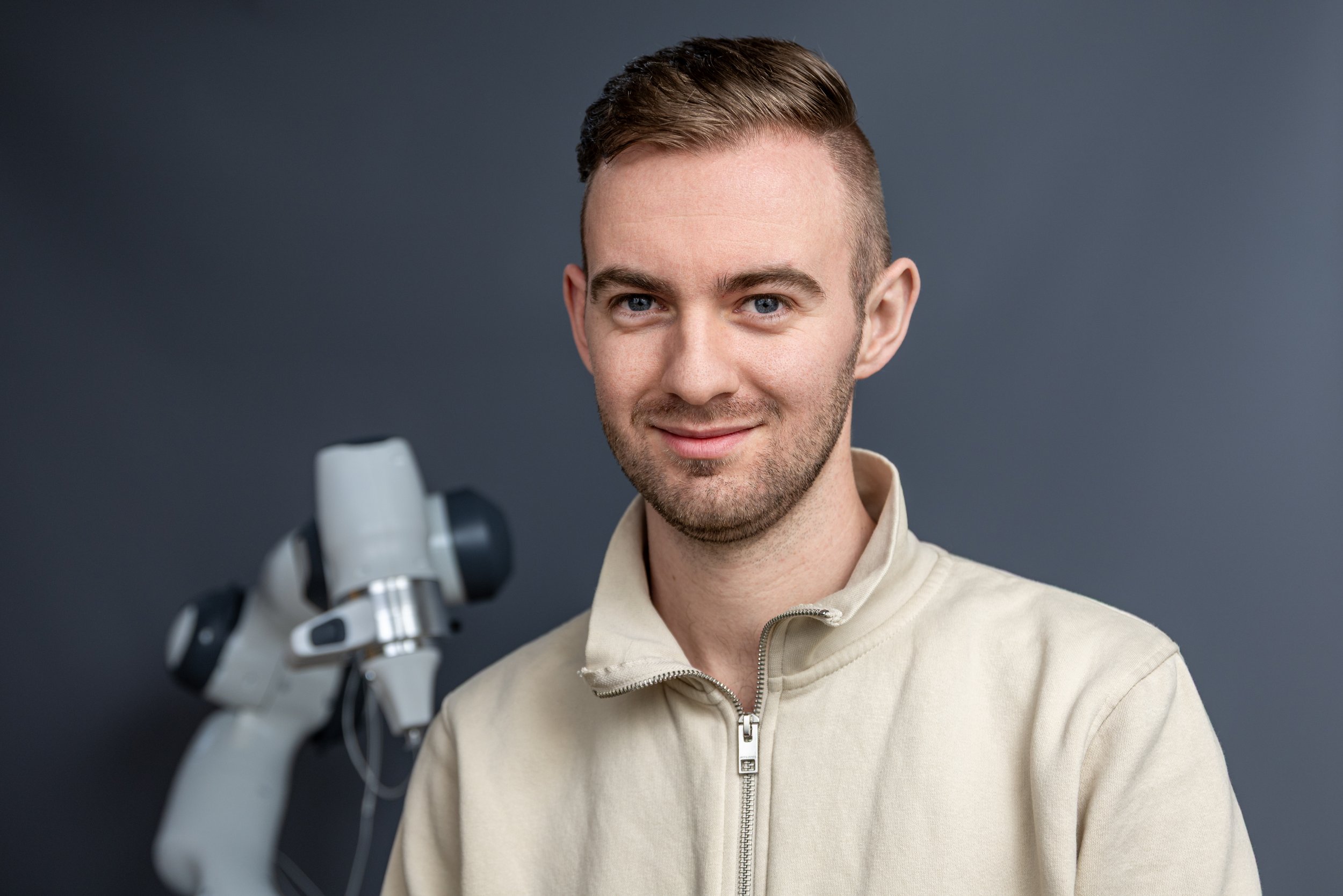 A young man with short brown hair and light stubble, smiling, standing next to a robotic arm, against a gray background.