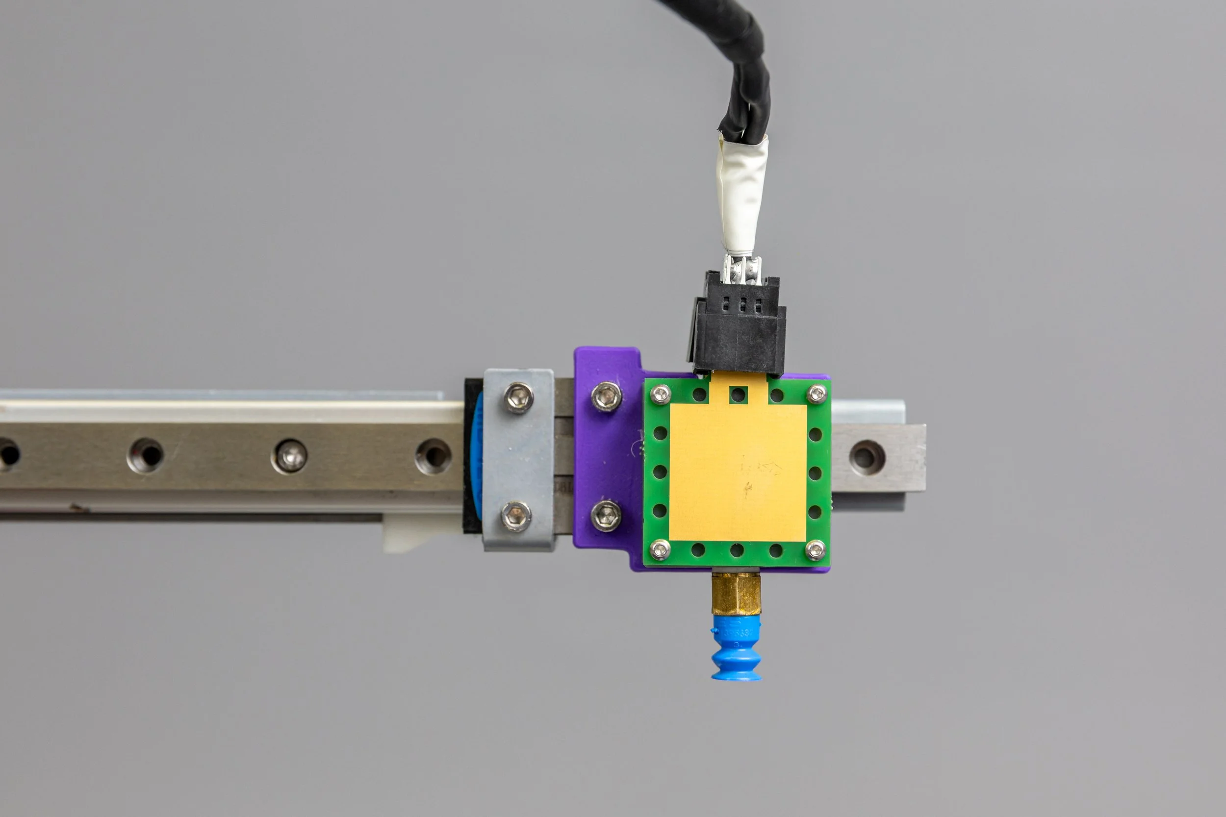 Close-up of a mechanical or robotic component mounted on a metal rail, with a yellow square in the center surrounded by green, purple, and blue elements, and an electrical cable attached on top.