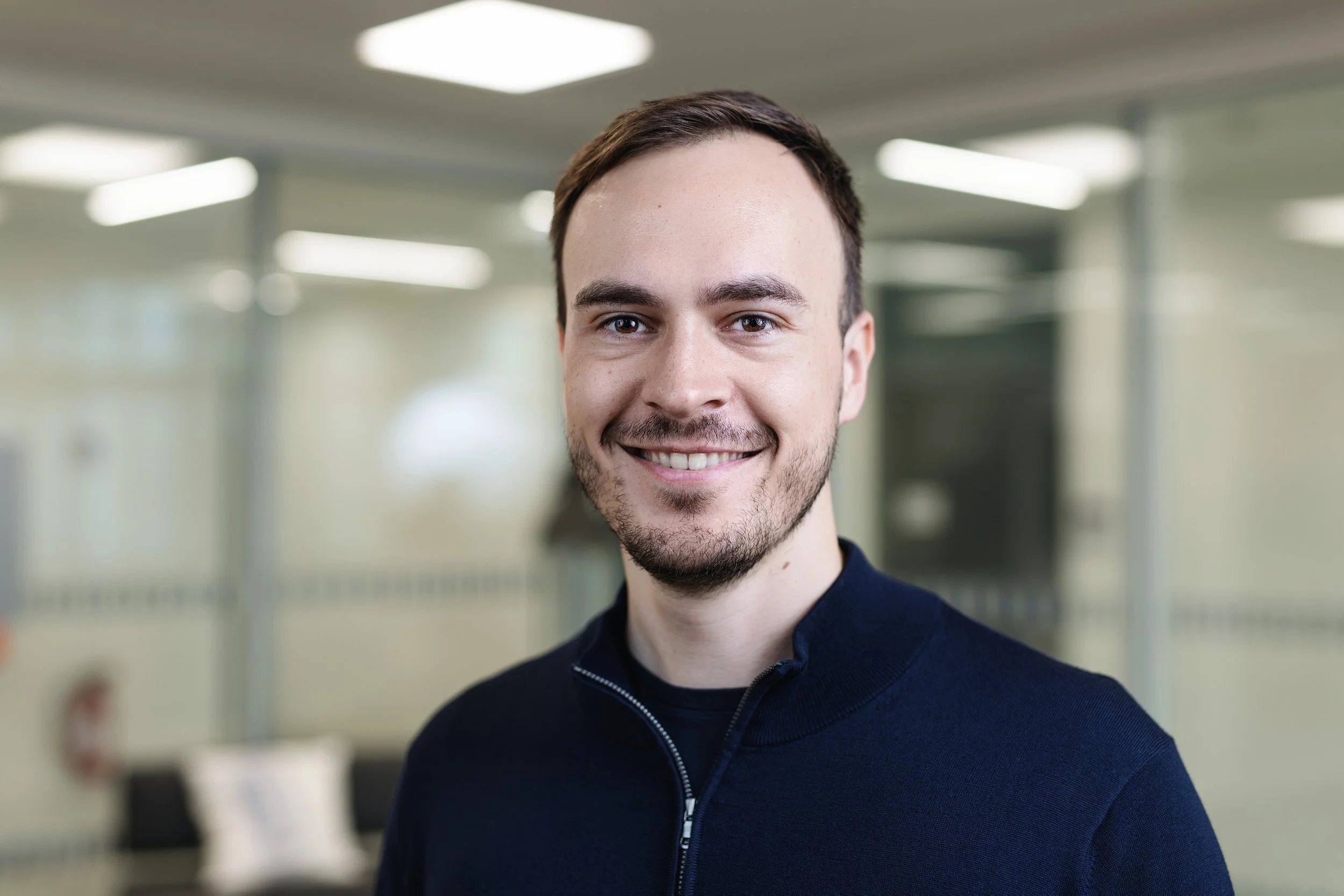 Ein lächelnder junger Mann mit kurzen braunen Haaren und Bart, der in einem Büro mit Glaswänden und heller Beleuchtung steht.