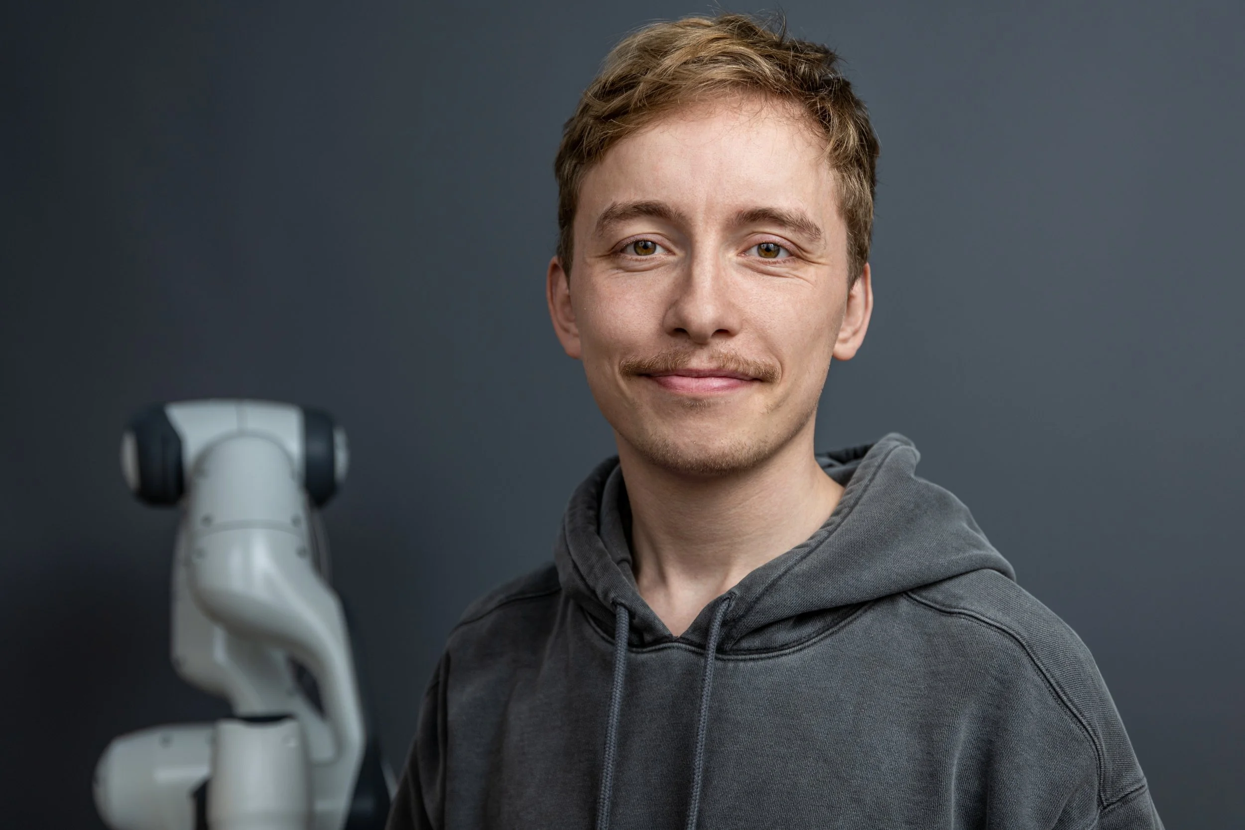 A young man with light brown hair and a mustache, wearing a gray hoodie, stands in front of a plain gray background with a robotic arm partially visible on the left side.