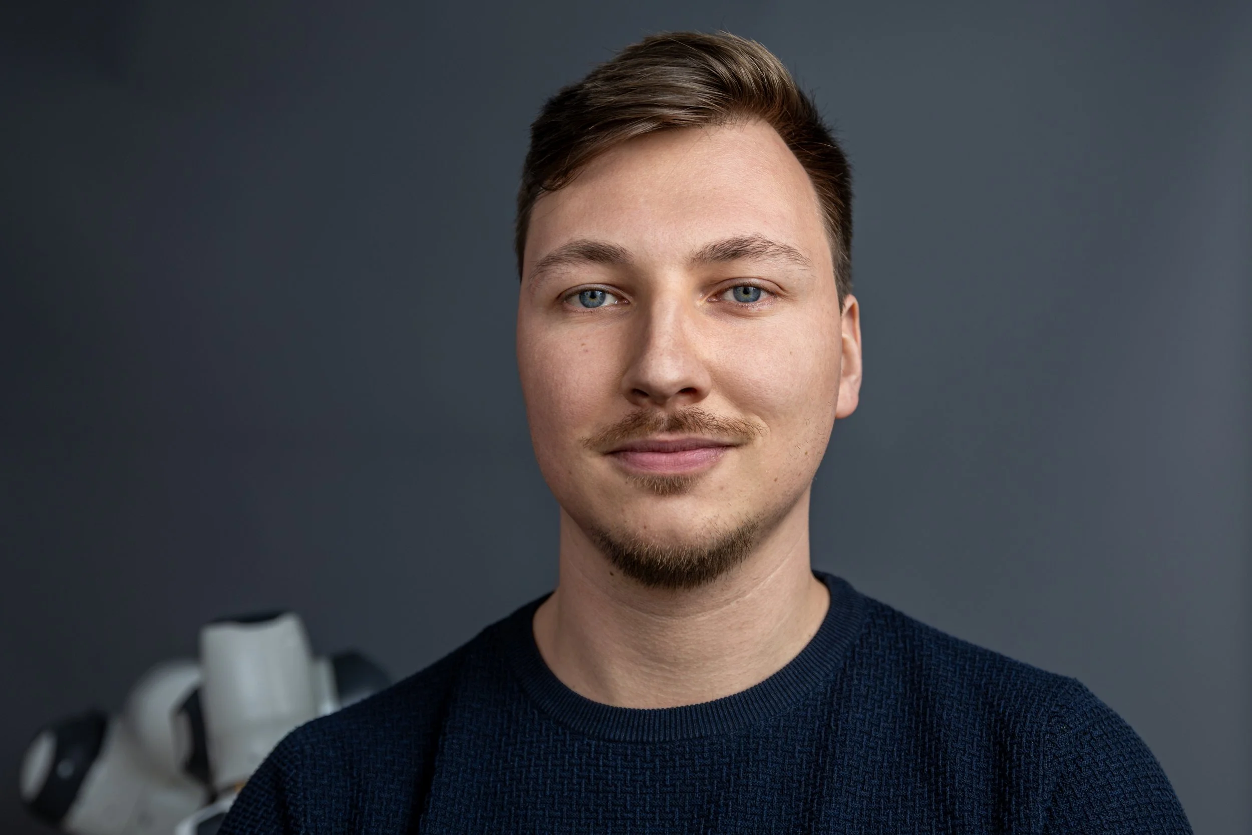 A young man with blue eyes and light brown hair styled to the side, smiling softly, wearing a dark blue sweater, against a plain dark gray background.