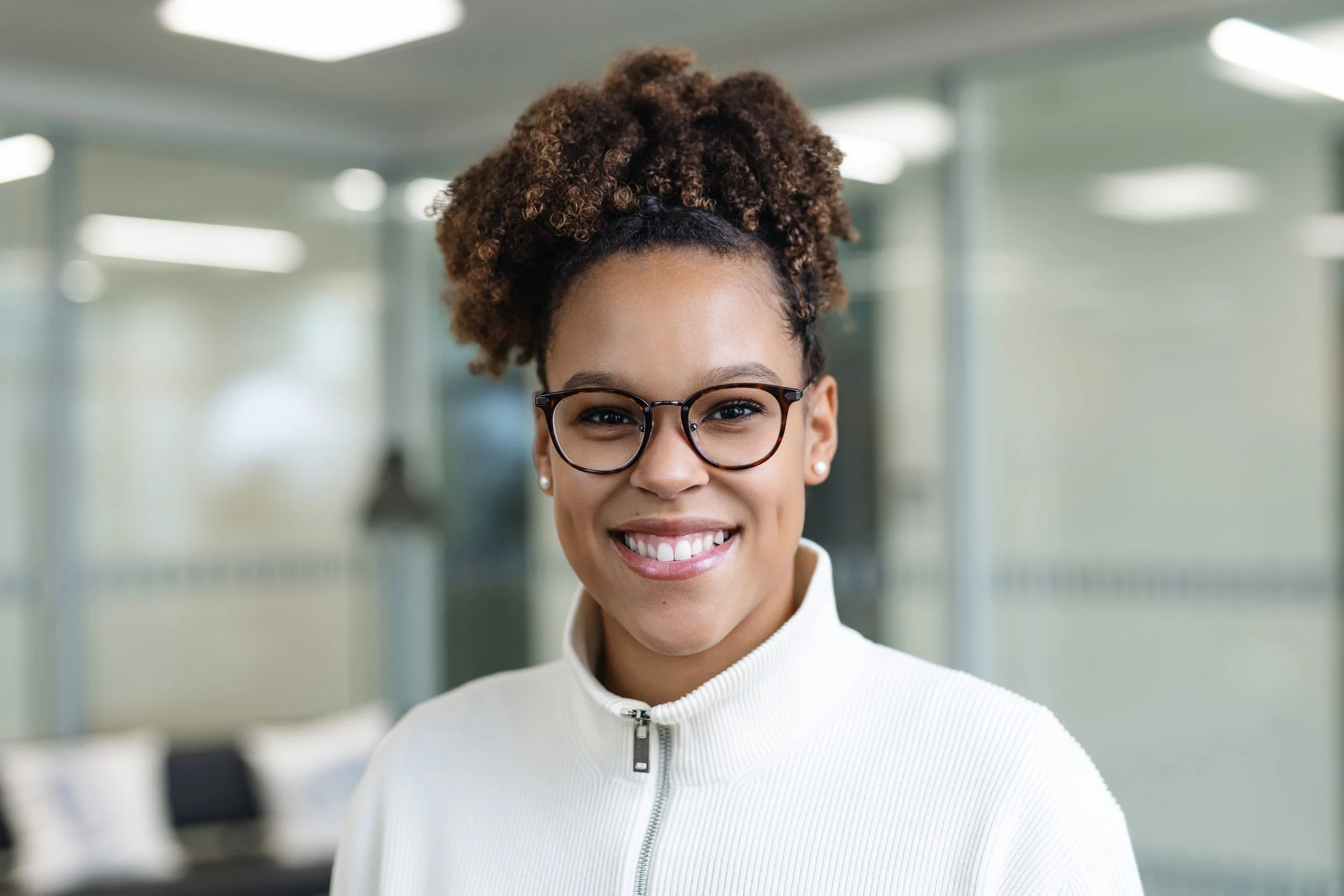 A woman smiling wearing glasses and a white zip-up jacket in a modern office setting.