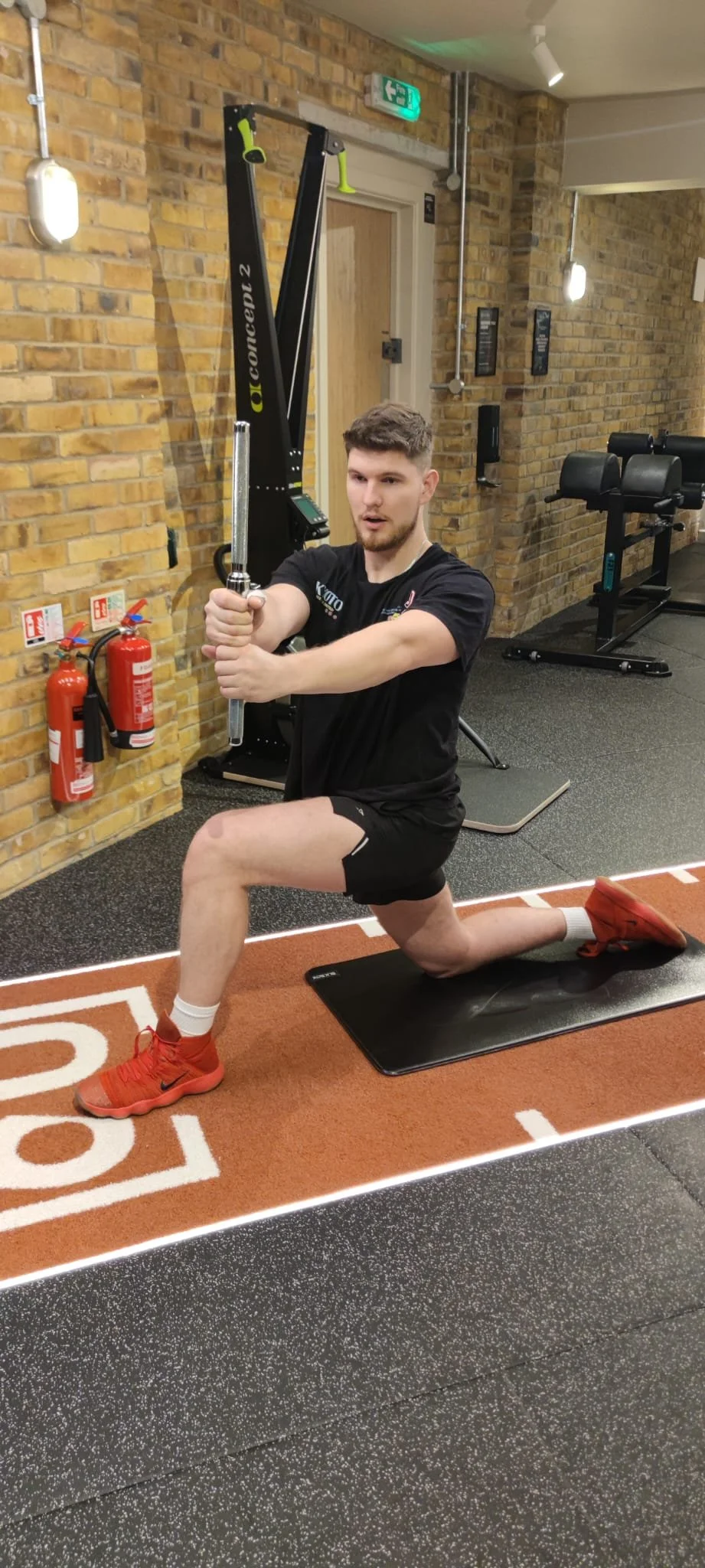 A young man with short brown hair, beard, and athletic build is performing a lunge exercise on a black mat in a gym. He is holding a barbell with weight plates at shoulder height, kneeling on one knee with the other foot planted flat on the ground. The gym has brick walls, exercise equipment, and fire extinguishers visible in the background.