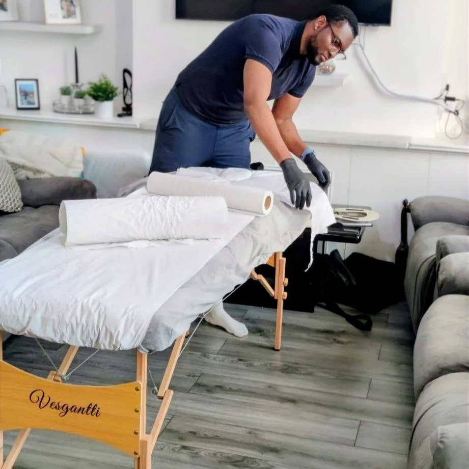 A man in a navy t-shirt and gloves preparing a massage table with white sheets and towels in a living room.