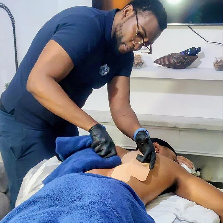 A patient is  lying on a treatment bed with a blue towel coveting his waist and legs, while a rehabilitation therapist is applying kinesiology tape on their back