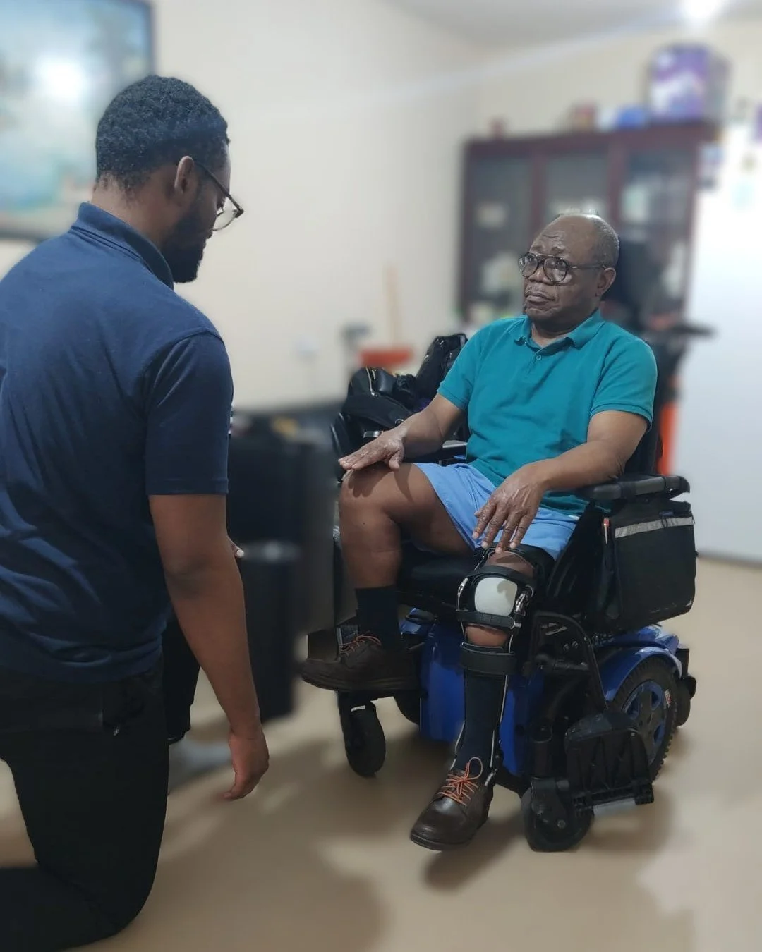 A rehabilotation therapist is kneeling in front of an a man in wheelchair supporting him through suided chair exercises