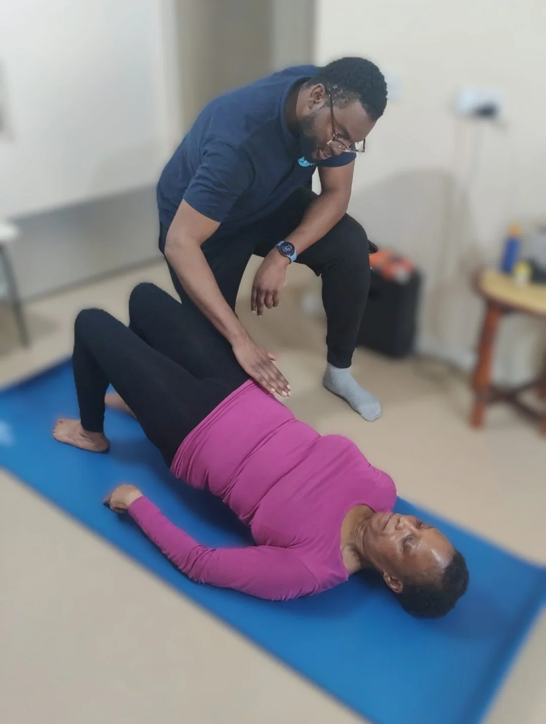 A rehabilitation therapits is assisting a woman doing a floor exercise on a blue yoga mat in a home setting
