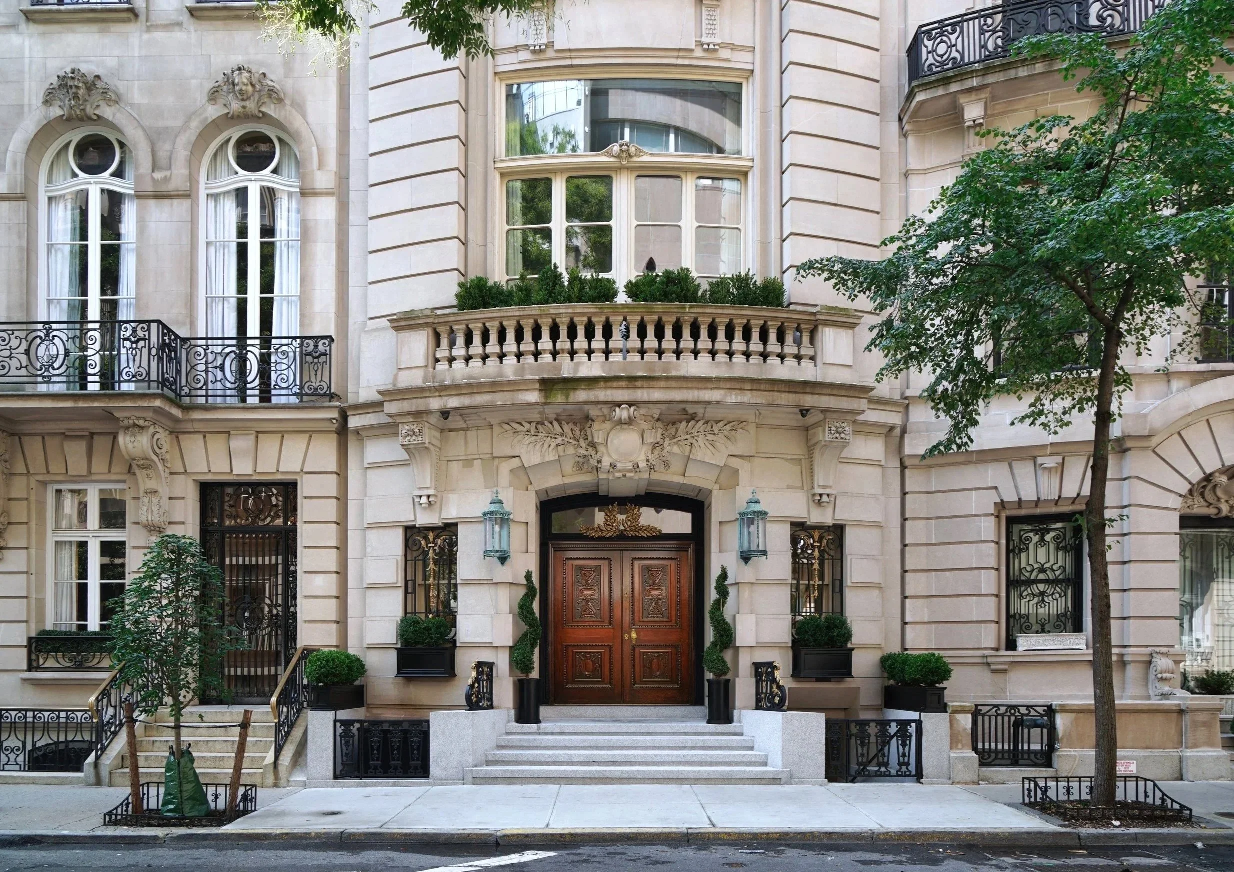 Facade of a beige stone building with ornate architectural details, large bay window, wooden front door, wrought iron balconies, and small plants around the entrance, flanked by trees.
