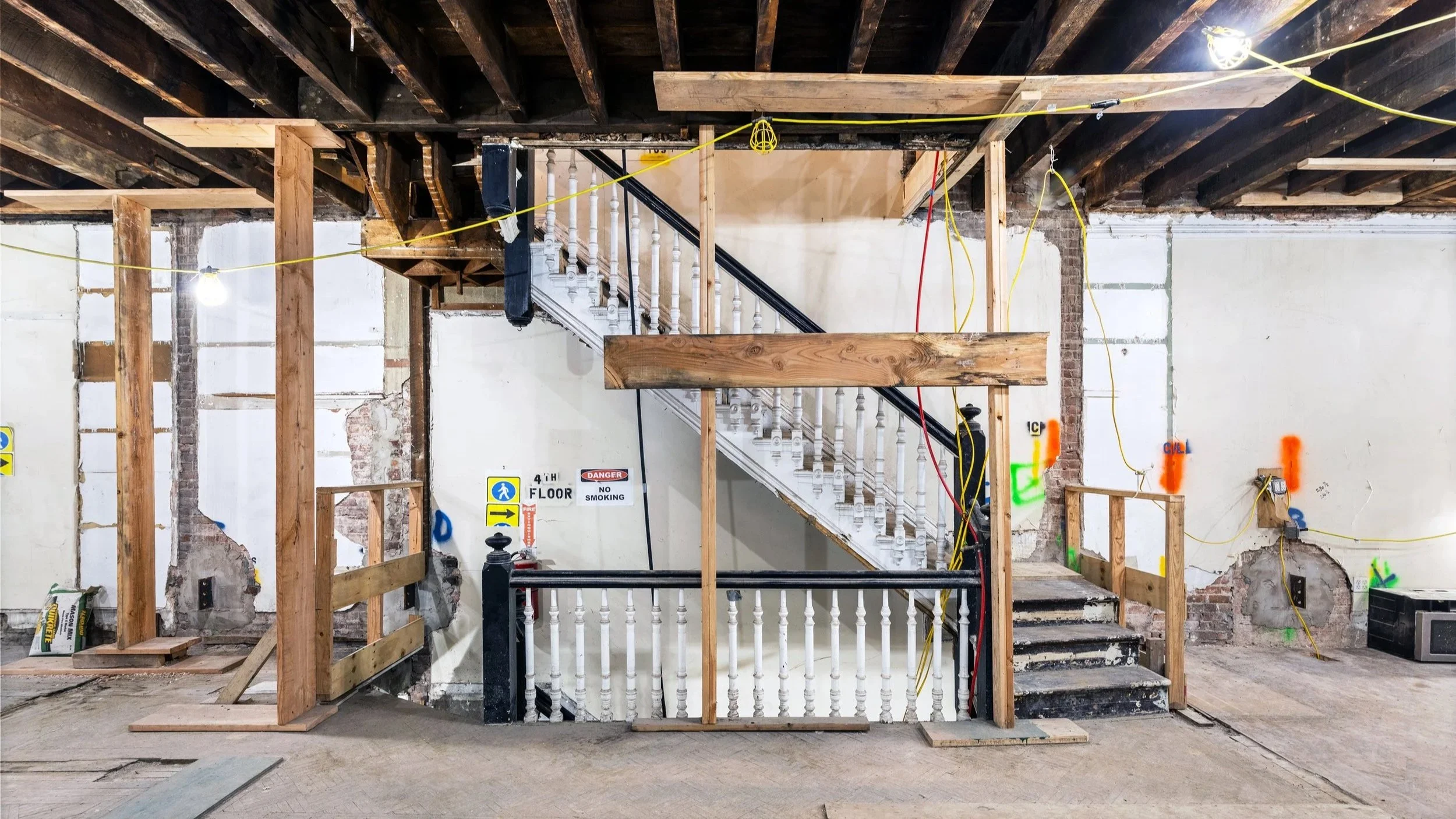 Interior of a building under renovation with exposed ceiling beams, construction lights, wooden framework, and a staircase with white balusters. Demolition of a Lenox Hill Townhouse- project development by Align Owner Represenation.