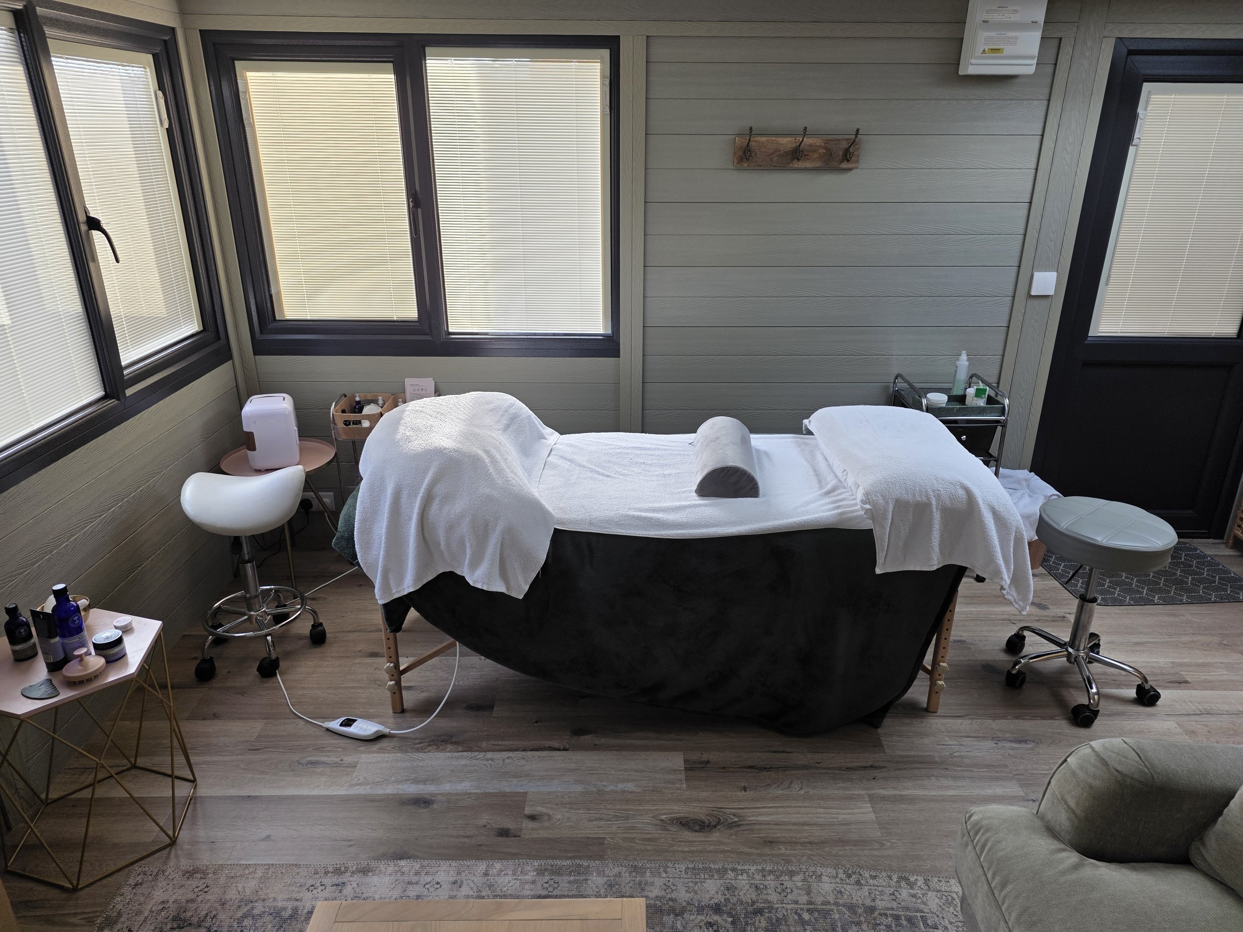 Spa or massage treatment table with pillow and blanket, located in a cozy room with wooden walls and windows, accented by warm candle and lantern lighting.