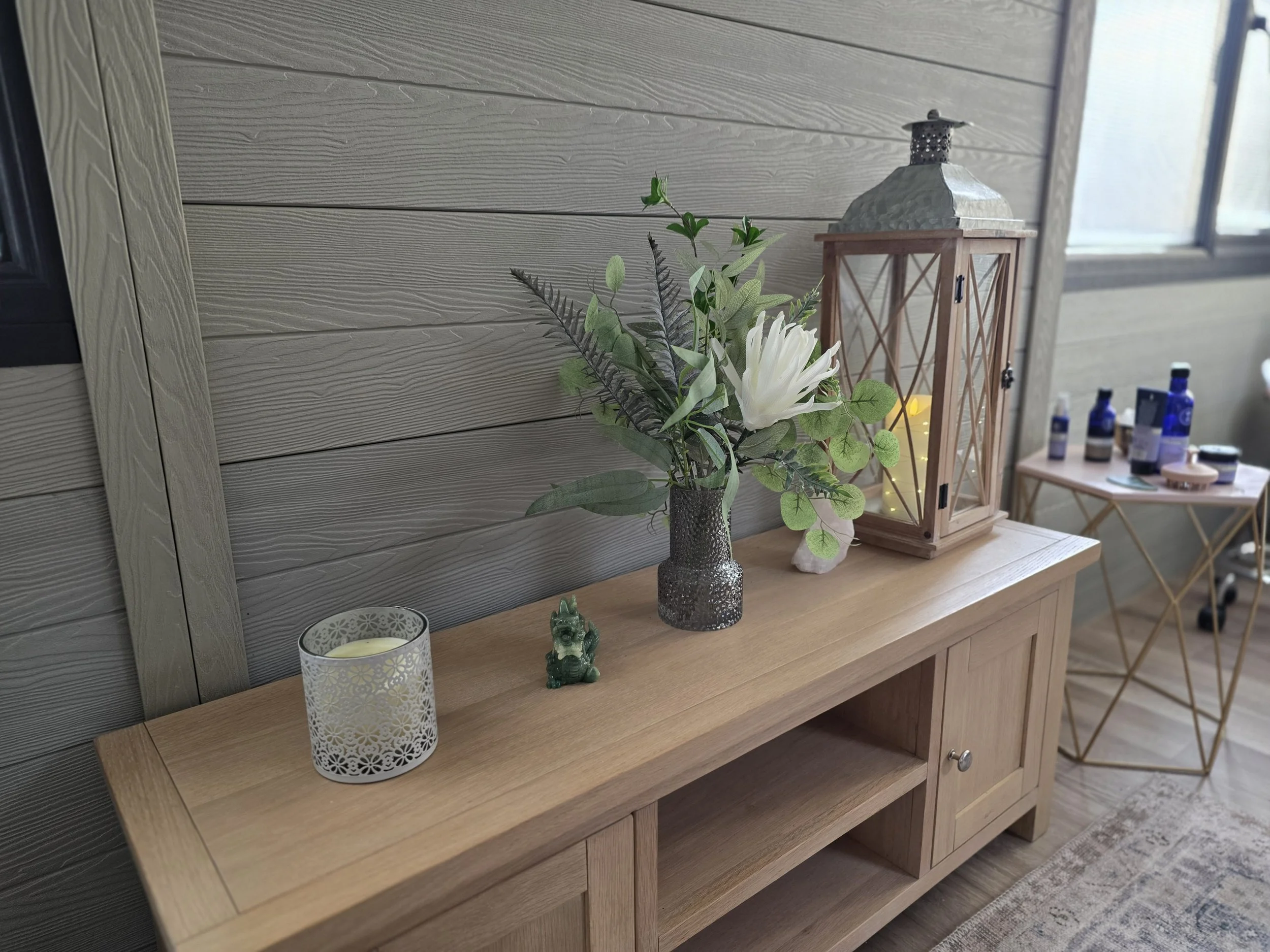 A decorative table arrangement featuring a lantern with a candle inside, a Buddha statue, a vase with white flowers and green foliage, and a table lamp with a black shade, all set against a wooden wall near a window.