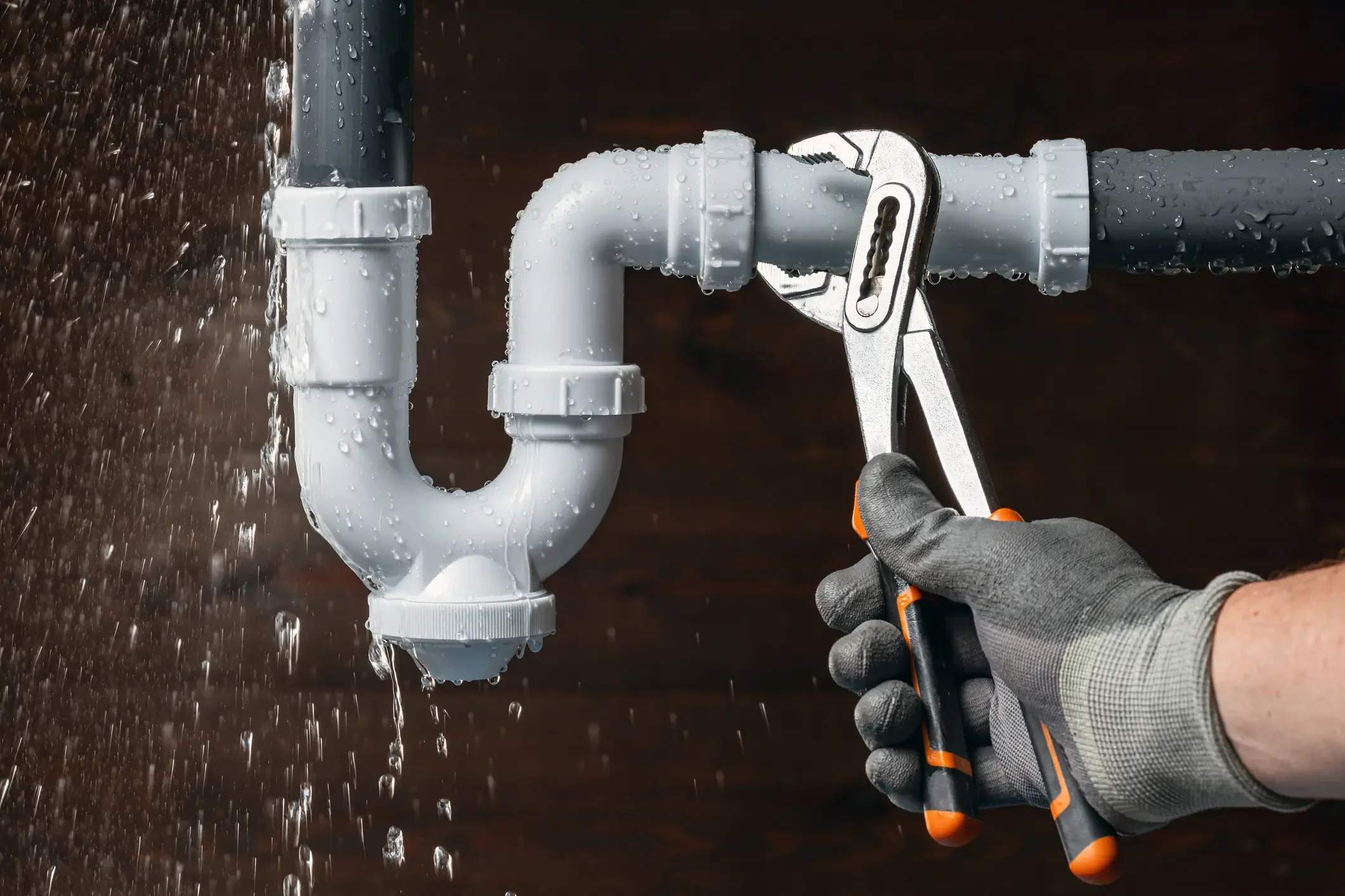 A close-up of a gloved hand using adjustable water pump pliers to tighten a white plastic P-trap drain pipe while water sprays out from a leak.