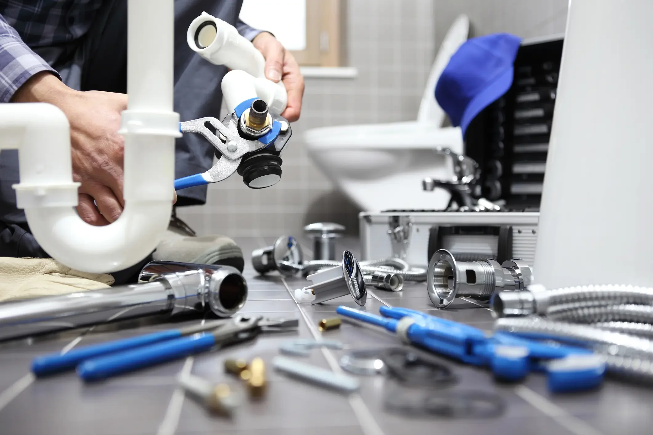 A plumber working on a bathroom floor surrounded by wrenches, hoses, and plumbing fittings, holding a pipe component near a toilet.