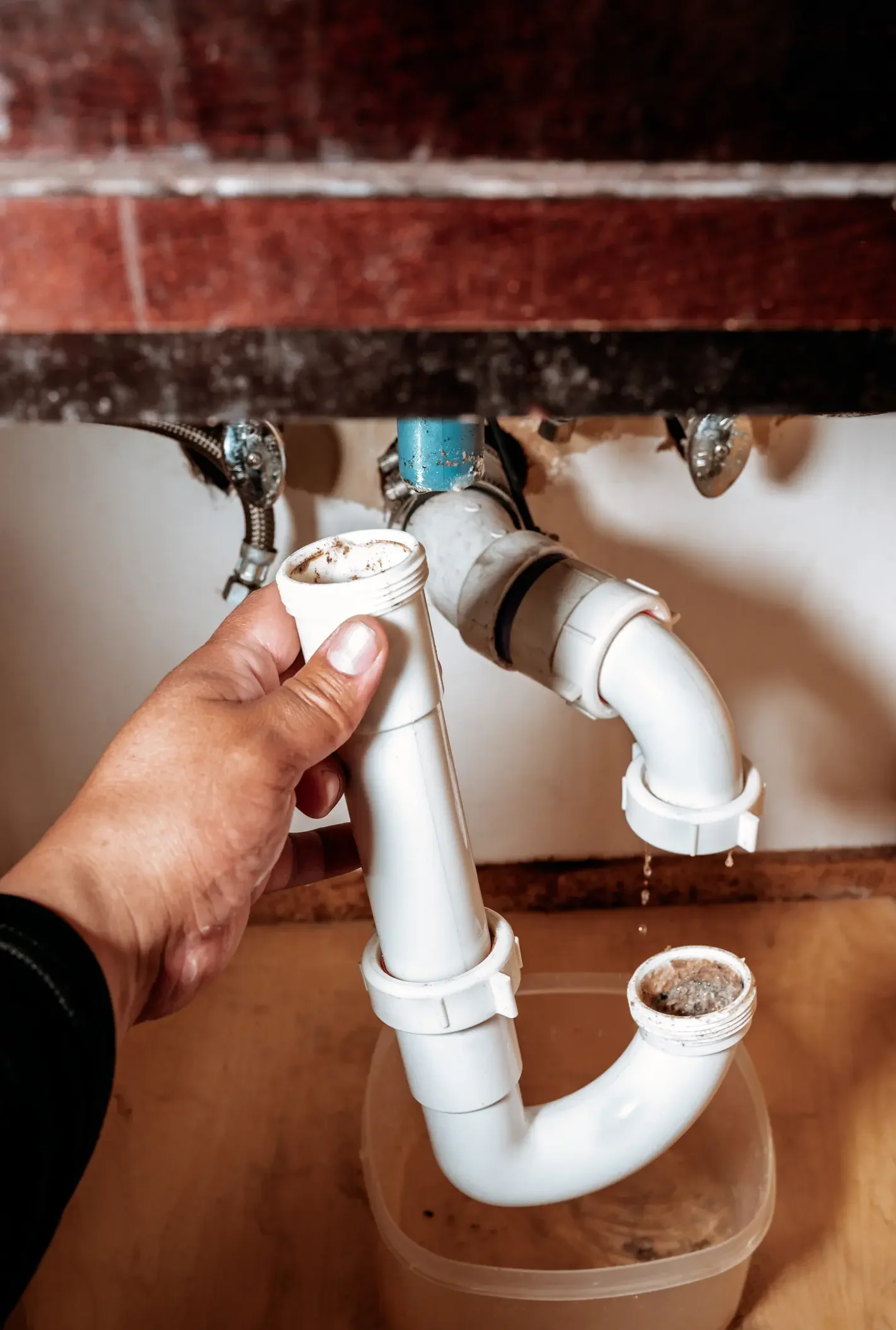 A hand holding a disassembled, dirty white plastic P-trap drain pipe under a sink with water dripping into a plastic container.