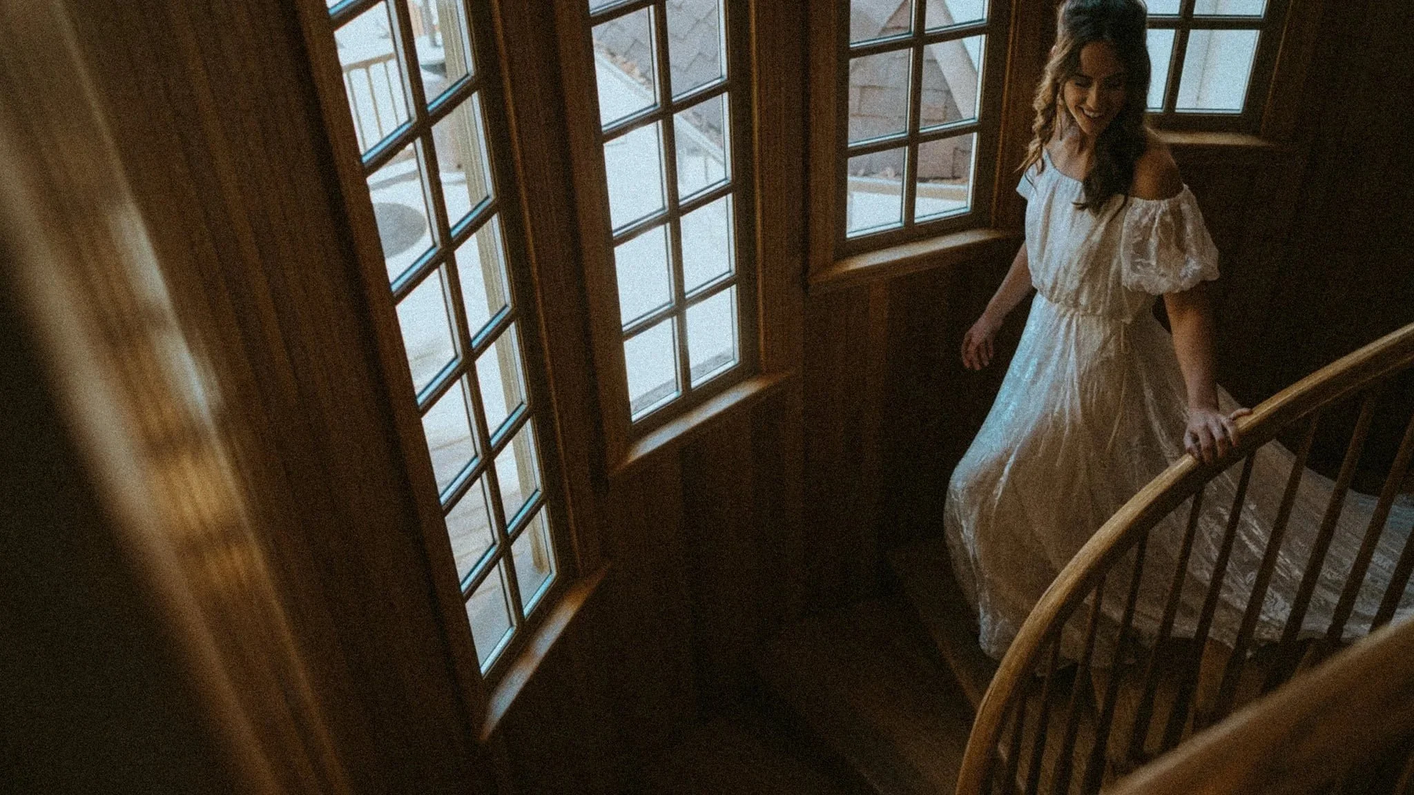 A woman in a white dress standing on a wooden staircase near a large window, smiling and looking down in Austin Texas