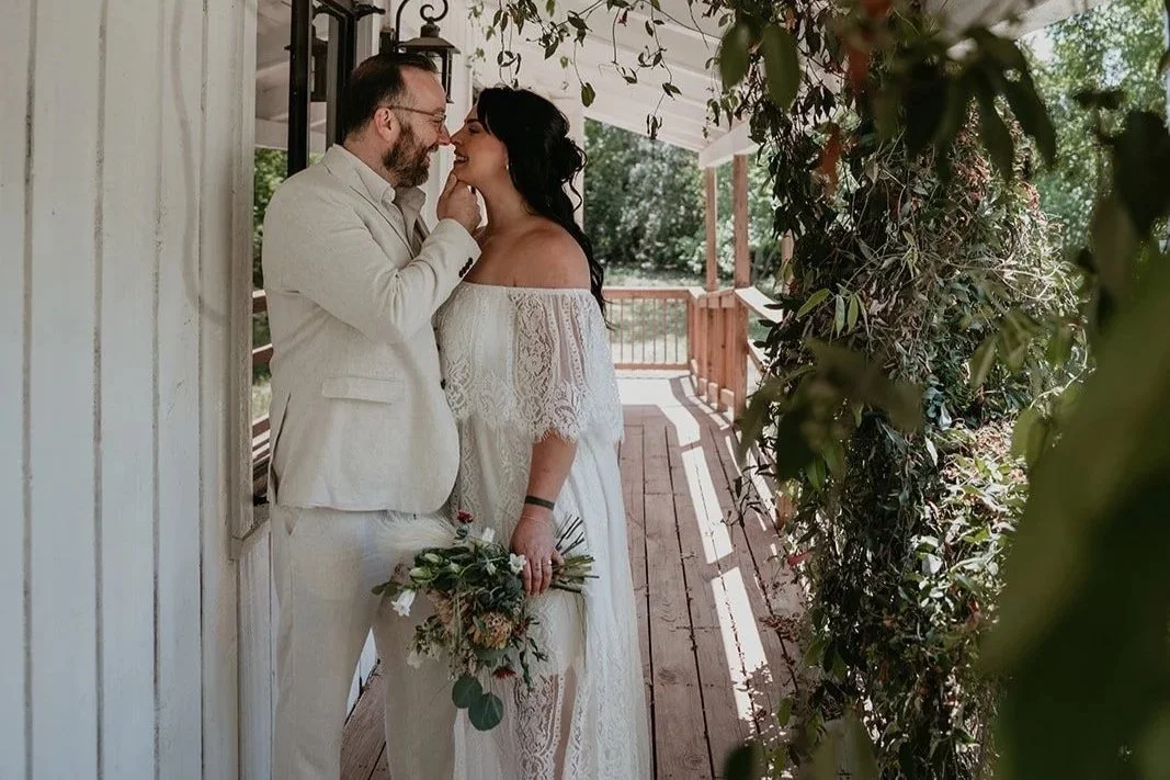 A couple dressed in white wedding attire standing close on a porch, smiling and touching noses, with the woman holding a bouquet of flowers and greenery, surrounded by hanging vines and foliage at an outdoor wedding venue in Austin Texas.