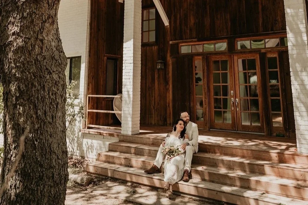 A couple sitting on wooden steps in front of a rustic house with white brick and wooden siding. The woman holds a bouquet of flowers, and the man sits close, with a tree in the foreground at an outdoor venue in Austin Texas.