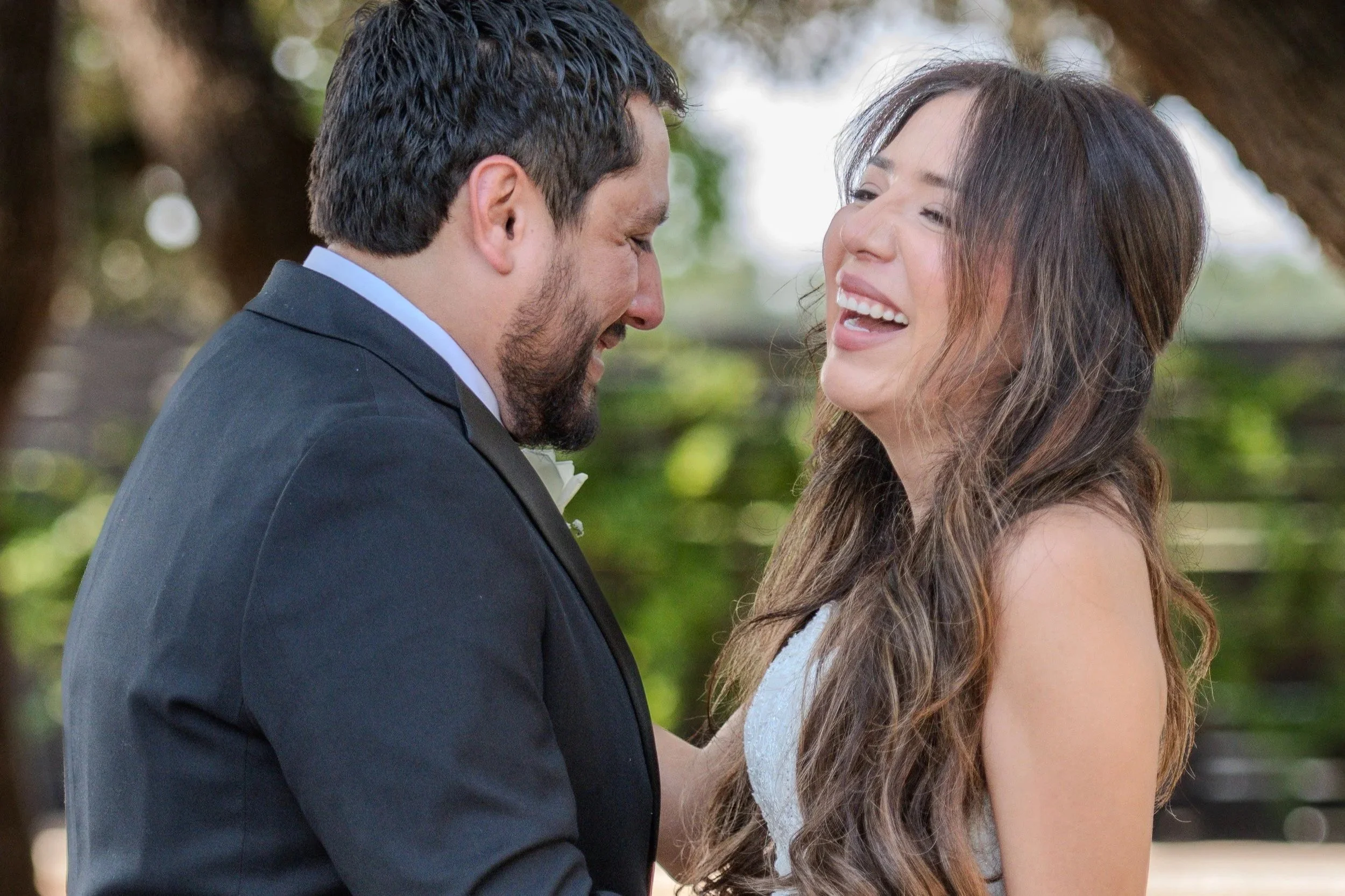 A man in a black tuxedo and a woman in a white dress share a joyful moment outdoors, smiling at each other.