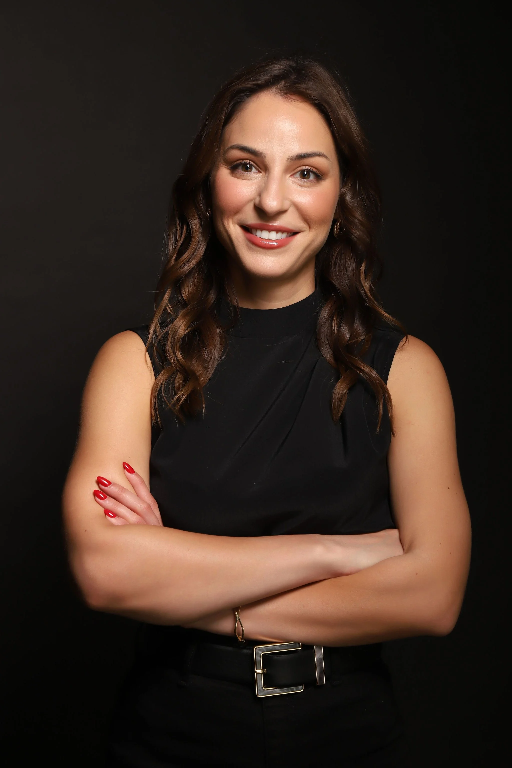 Portrait of a woman with wavy brown hair wearing a black sleeveless top, arms crossed, smiling, against a dark background.