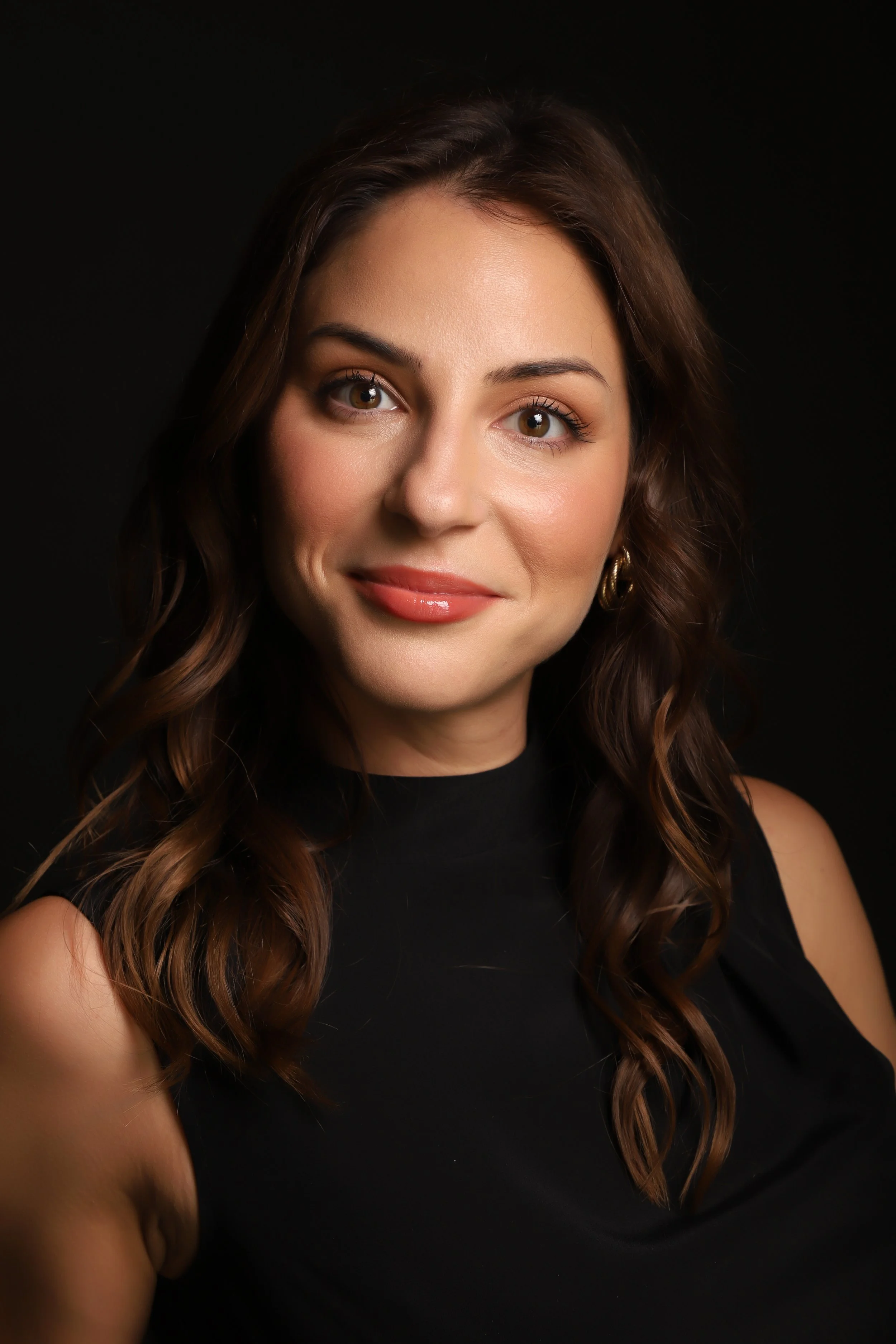 Portrait of a woman with wavy brown hair, wearing a black sleeveless top, with a dark background.
