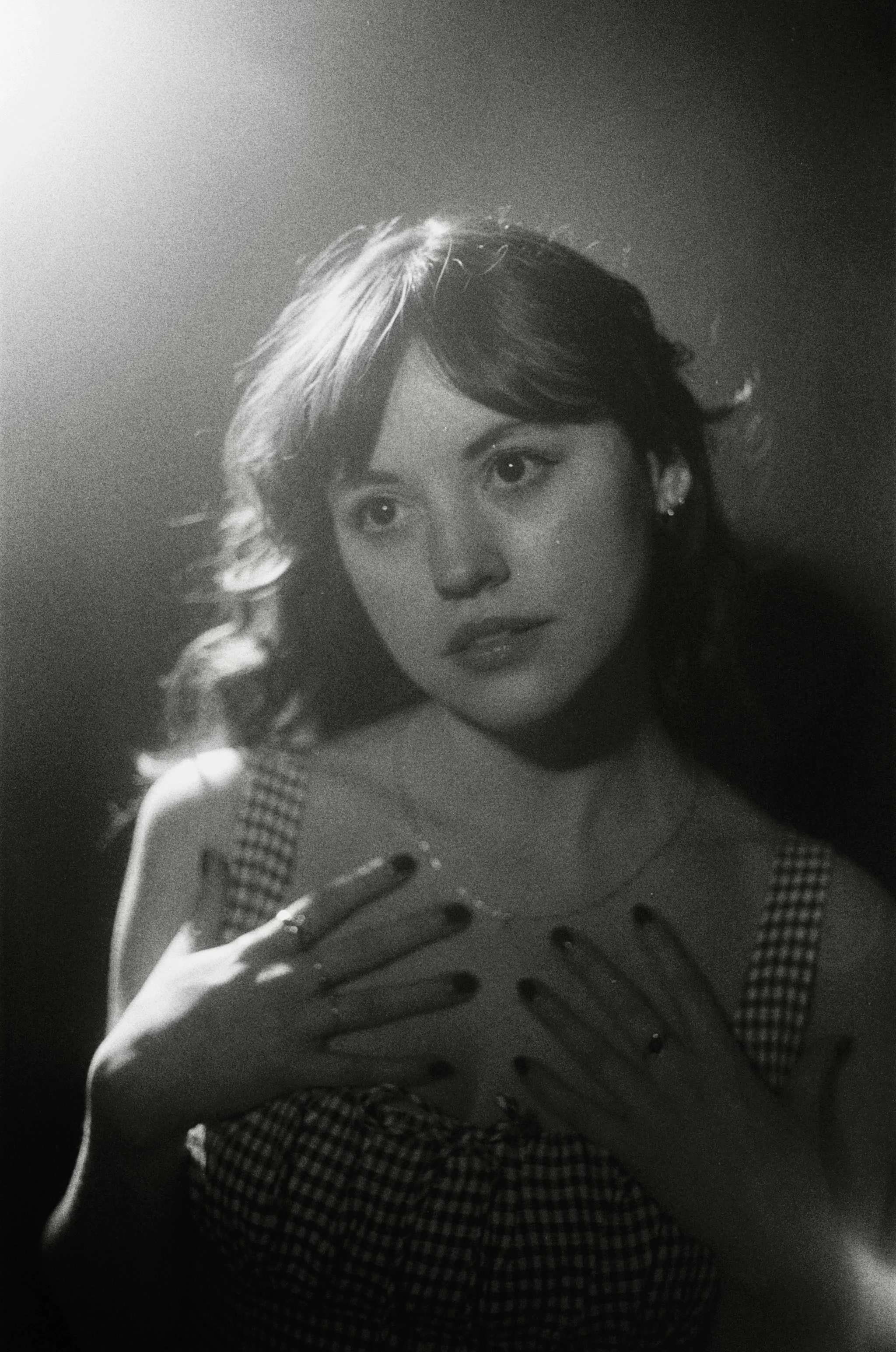 A black and white portrait of a young woman with wavy hair, wearing a checkered sleeveless top, looking to her left, with her hands placed on her chest and a spotlight illuminating her face.