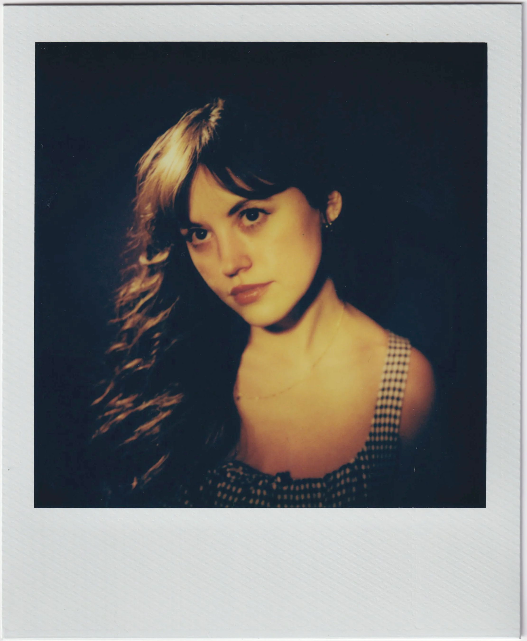 A young woman with long wavy hair, wearing a checkered dress, and a delicate necklace, posing against a dark background.