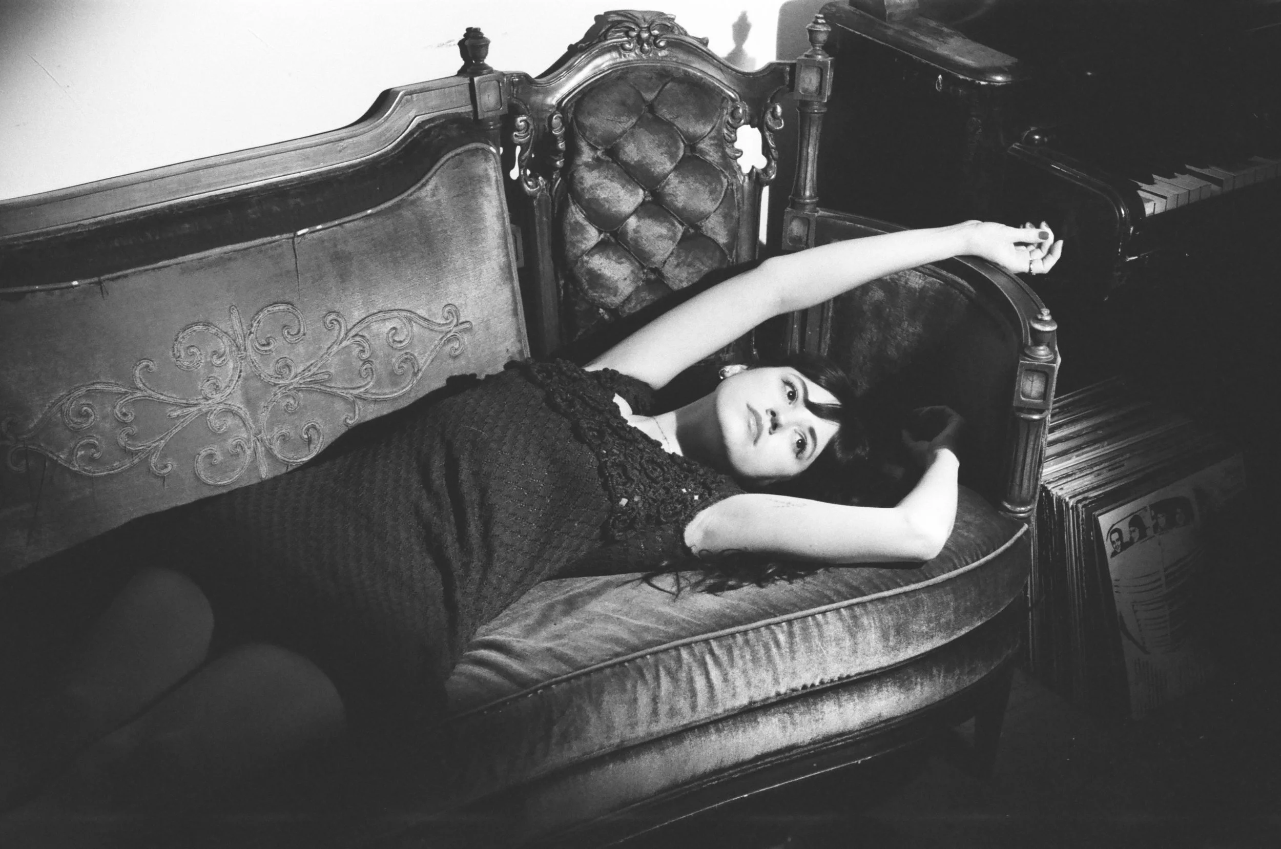 A young woman with dark hair and bangs lying on an ornate vintage sofa with her arms stretched above her head. She is wearing a dark, textured dress and resting her head on a cushion. A piano and vinyl records are visible in the background, and the p