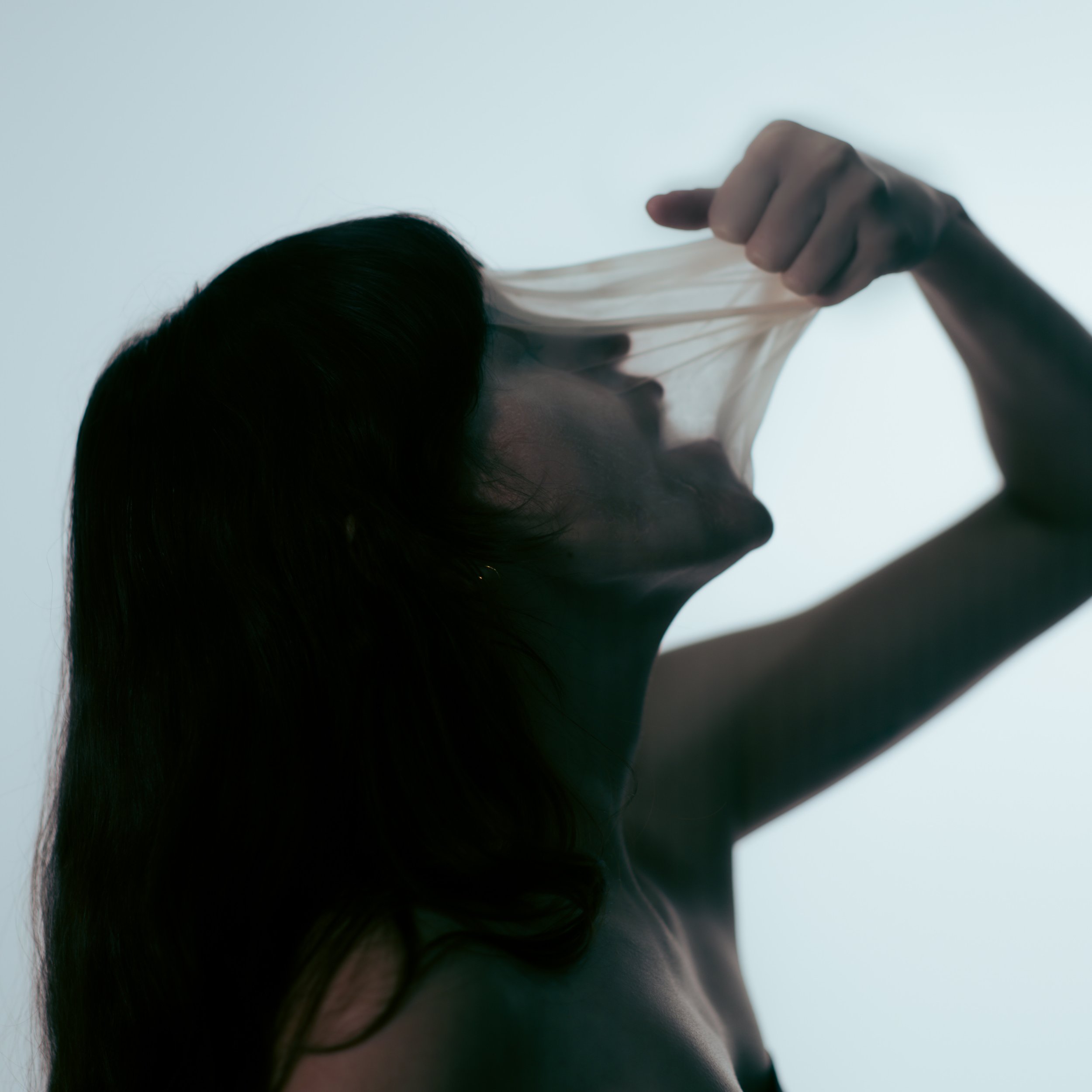 Silhouette of a woman pulling a sheer fabric over her face against a pale background.