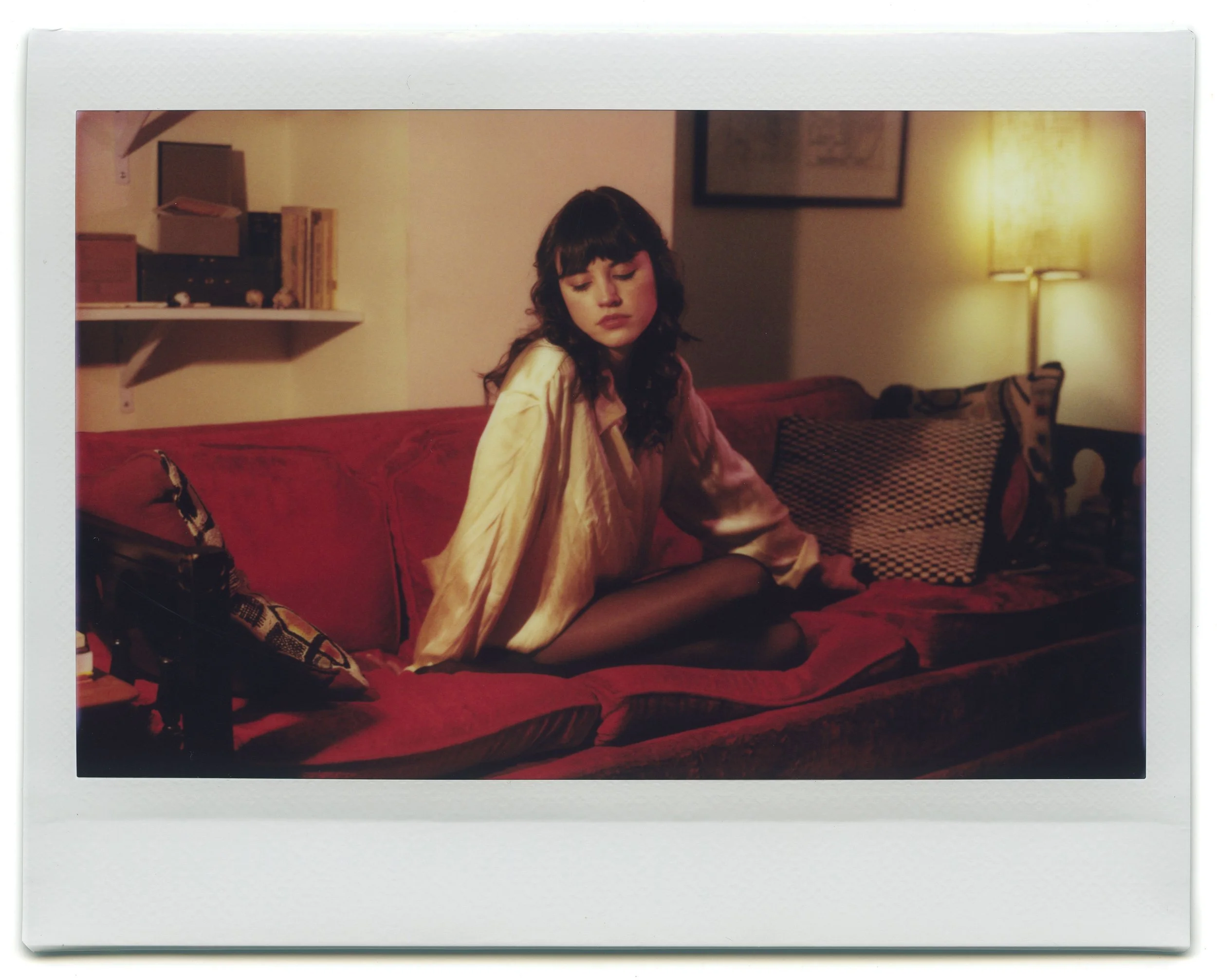 A woman with dark curly hair wearing a satin silk cream-colored shirt, sitting on a red velvet couch with her eyes closed and her head tilted downward.