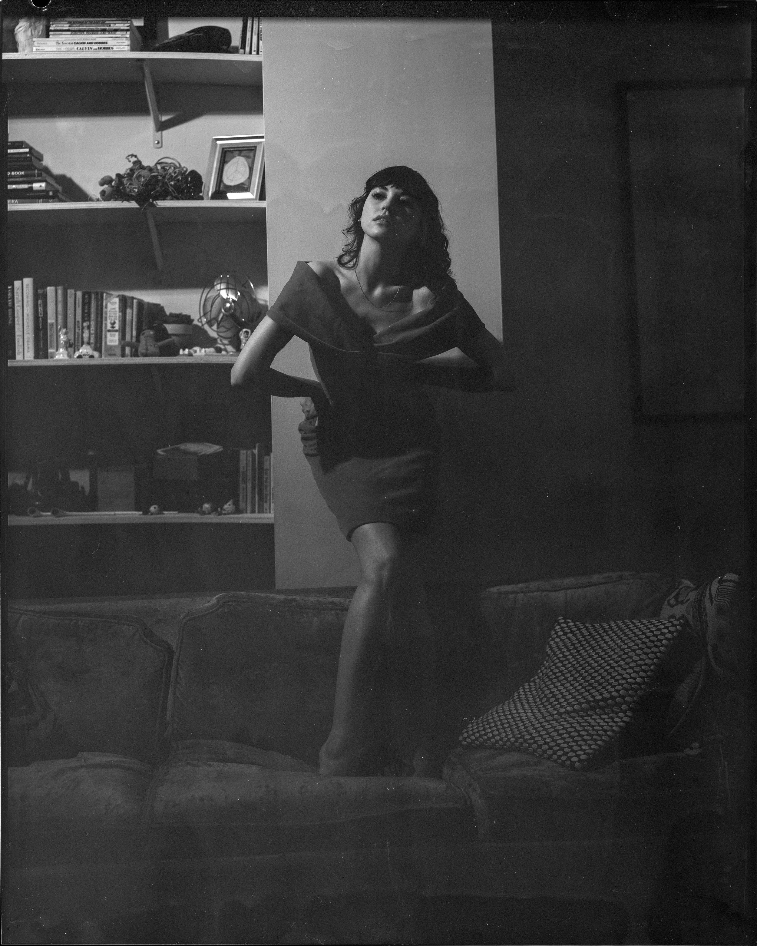 A black and white photo of a woman standing on a vintage sofa in a room with bookshelves and decorated with framed photographs and objects.