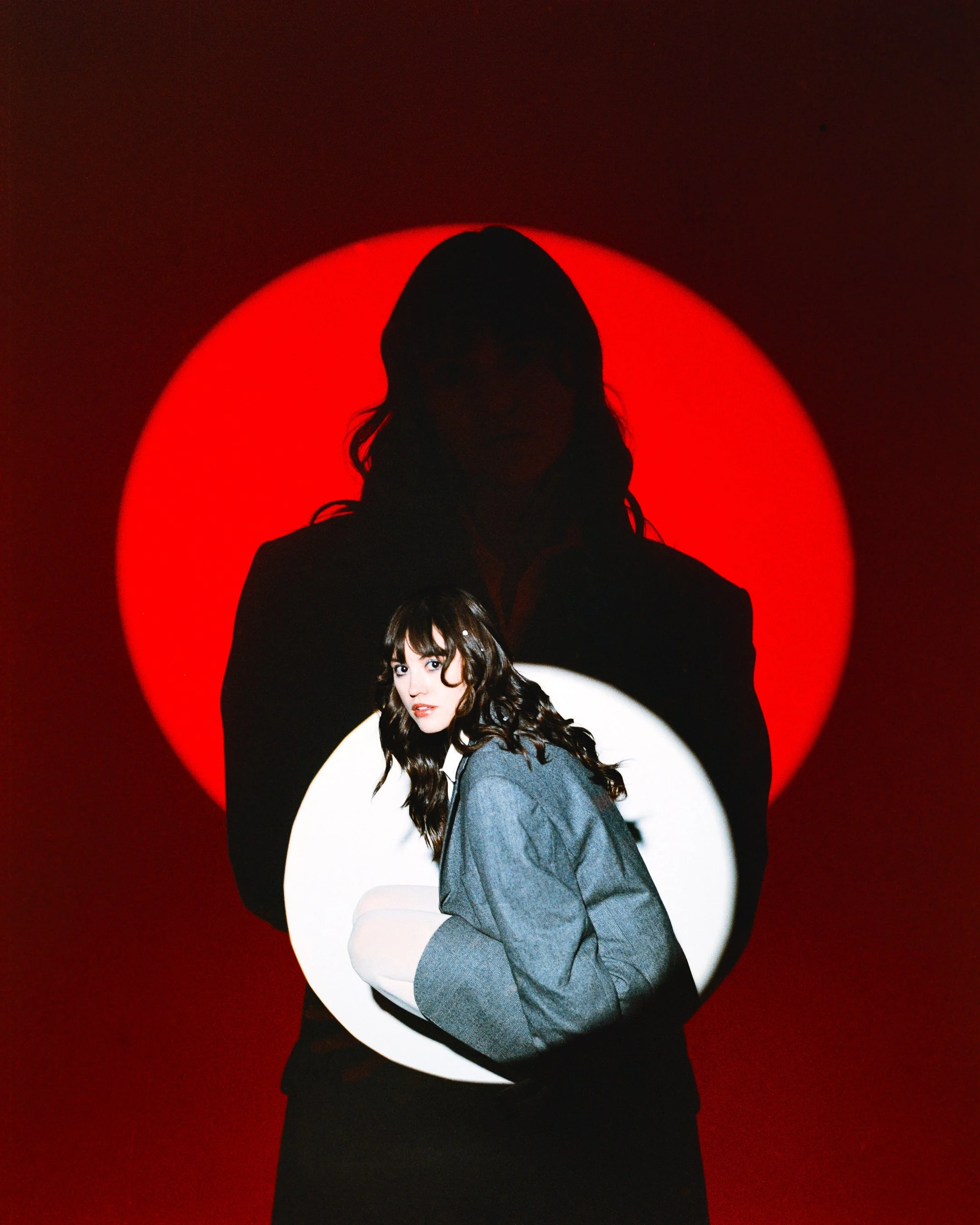 A woman with long wavy hair and a light-colored jacket, sitting in front of a red background with a white circle, and a shadow of another woman behind her.