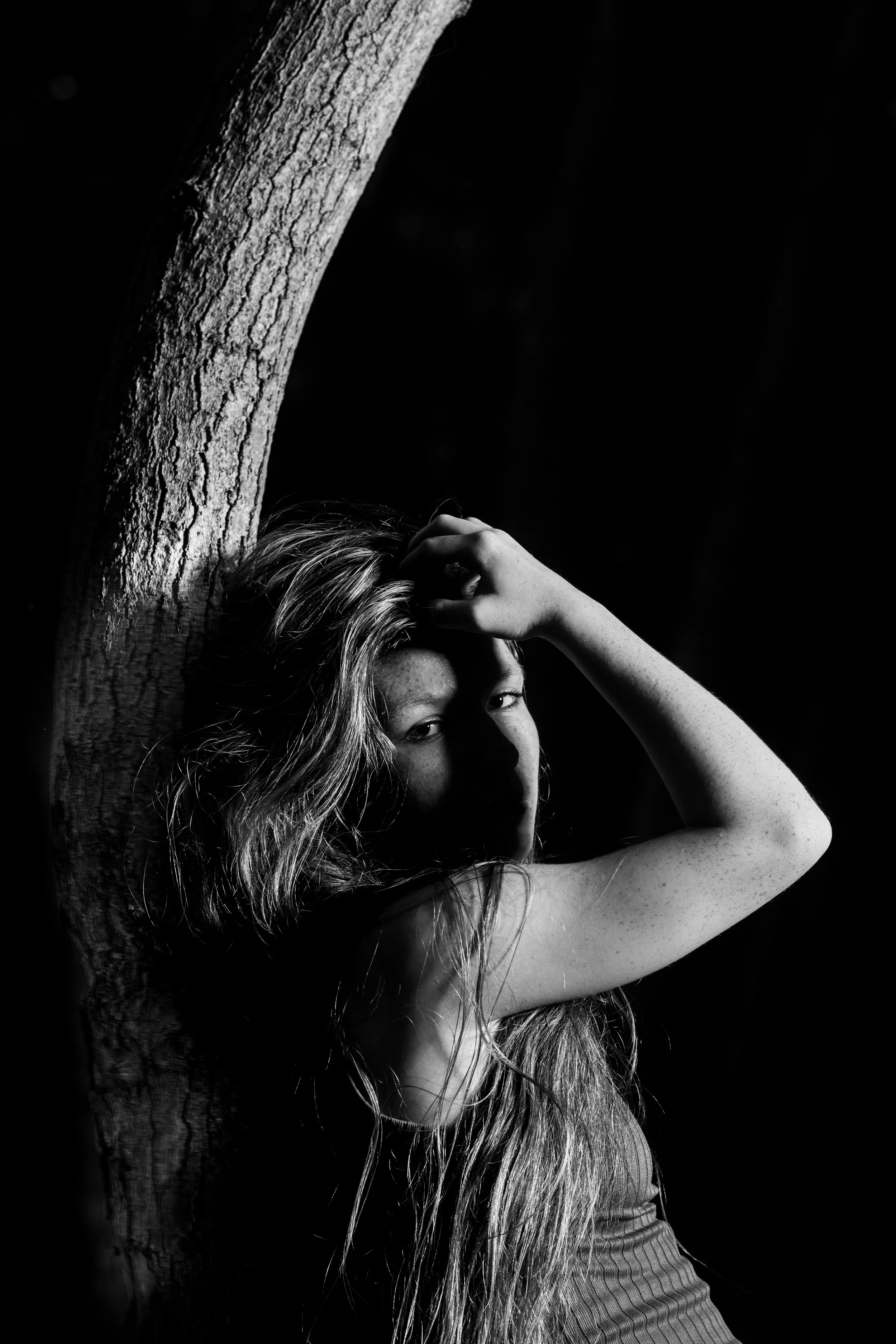 Black and white photograph of a woman with long, wavy hair leaning against a tree, with one hand resting on her forehead and a shadow partially obscuring her face.