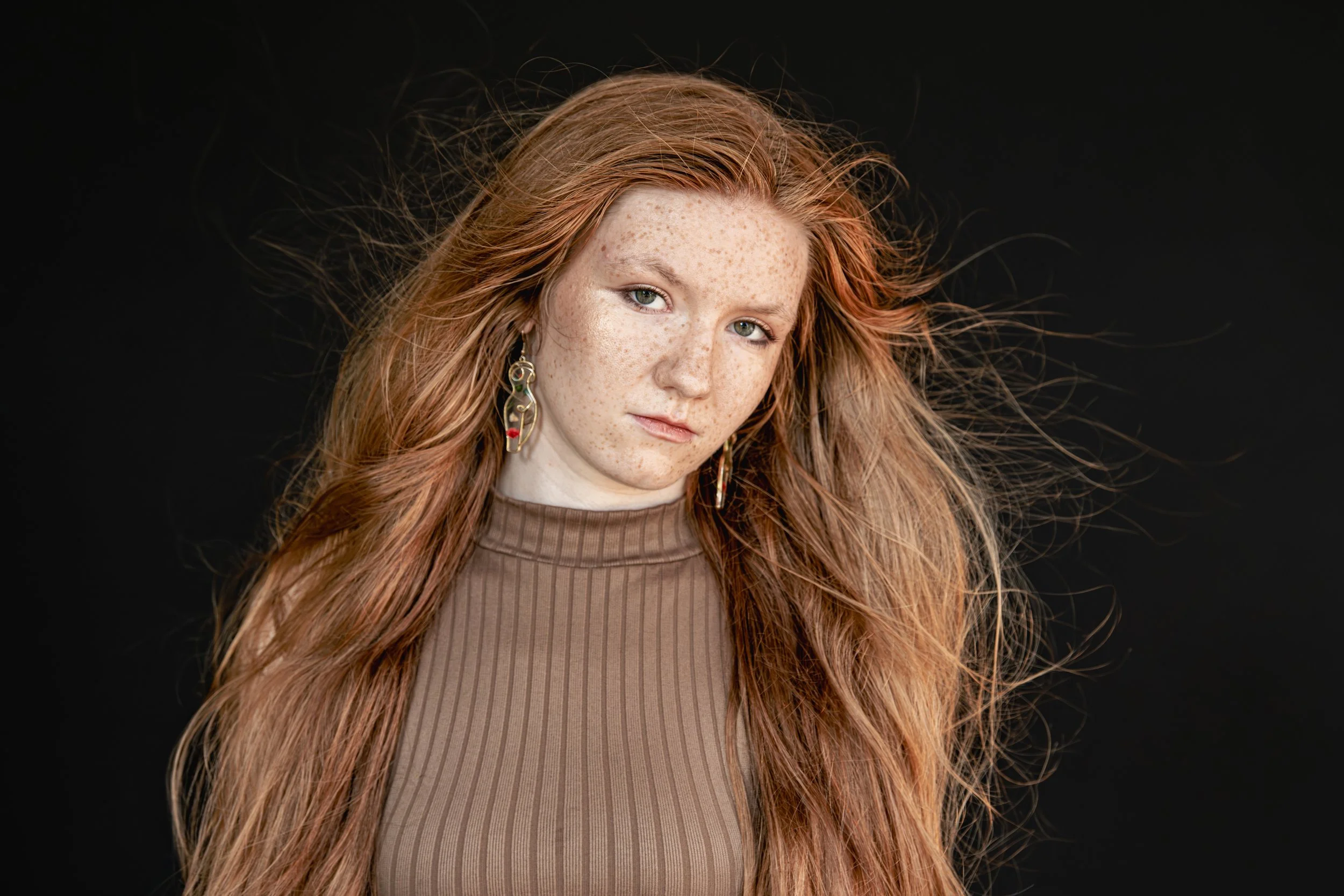 A portrait of a young woman with red hair, fair skin with freckles, wearing a brown ribbed top and colorful dangling earrings, against a black background.