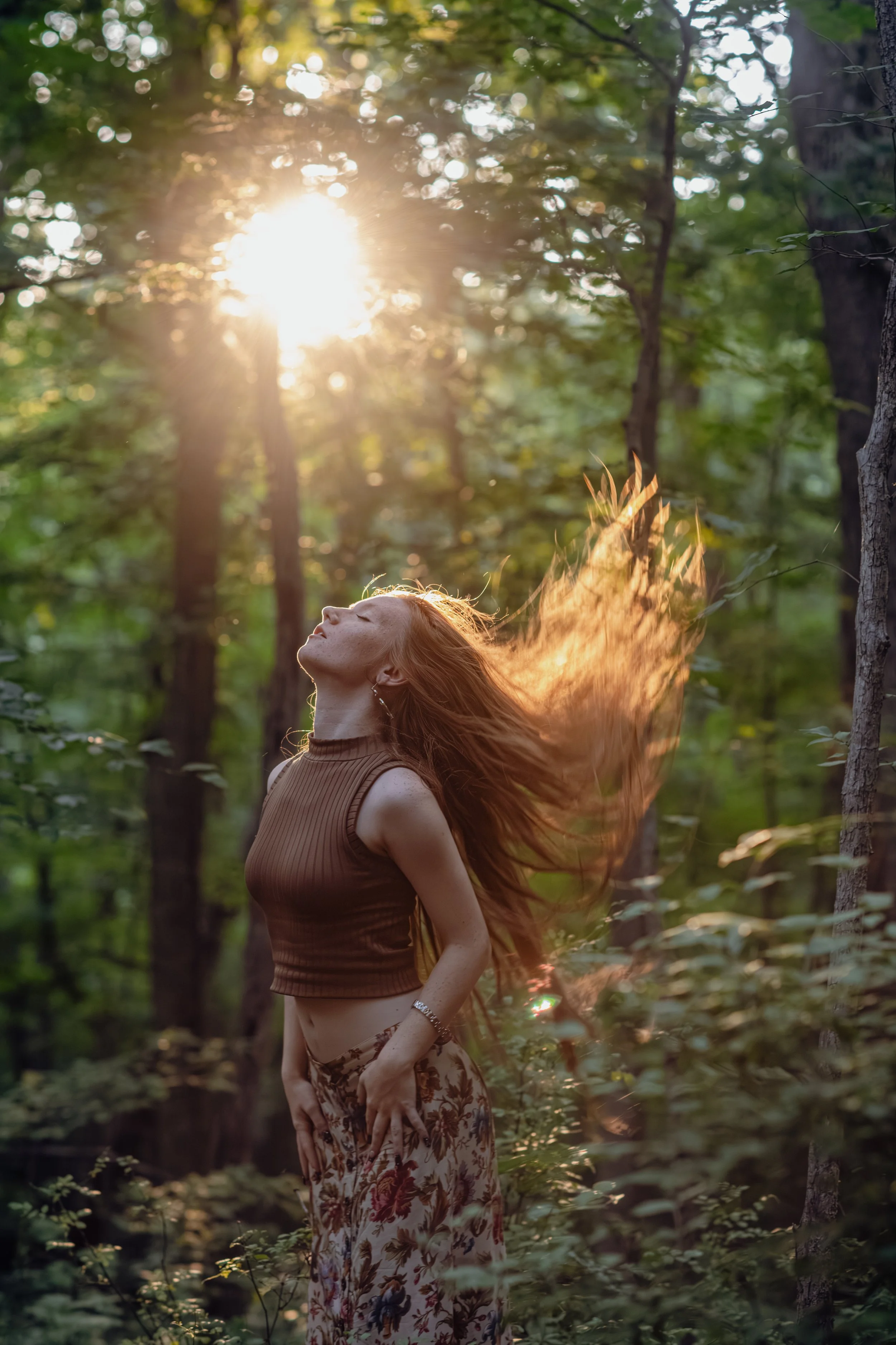 A woman with long red hair standing in a forest with sunlight shining through the trees, her eyes closed, with her hair flowing behind her.