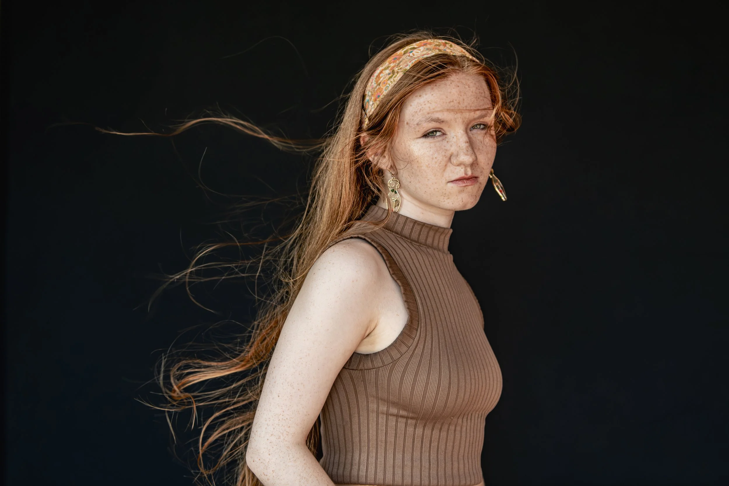 A young woman with long, red hair and freckles, wearing a brown sleeveless turtleneck top and floral headband, against a black background.
