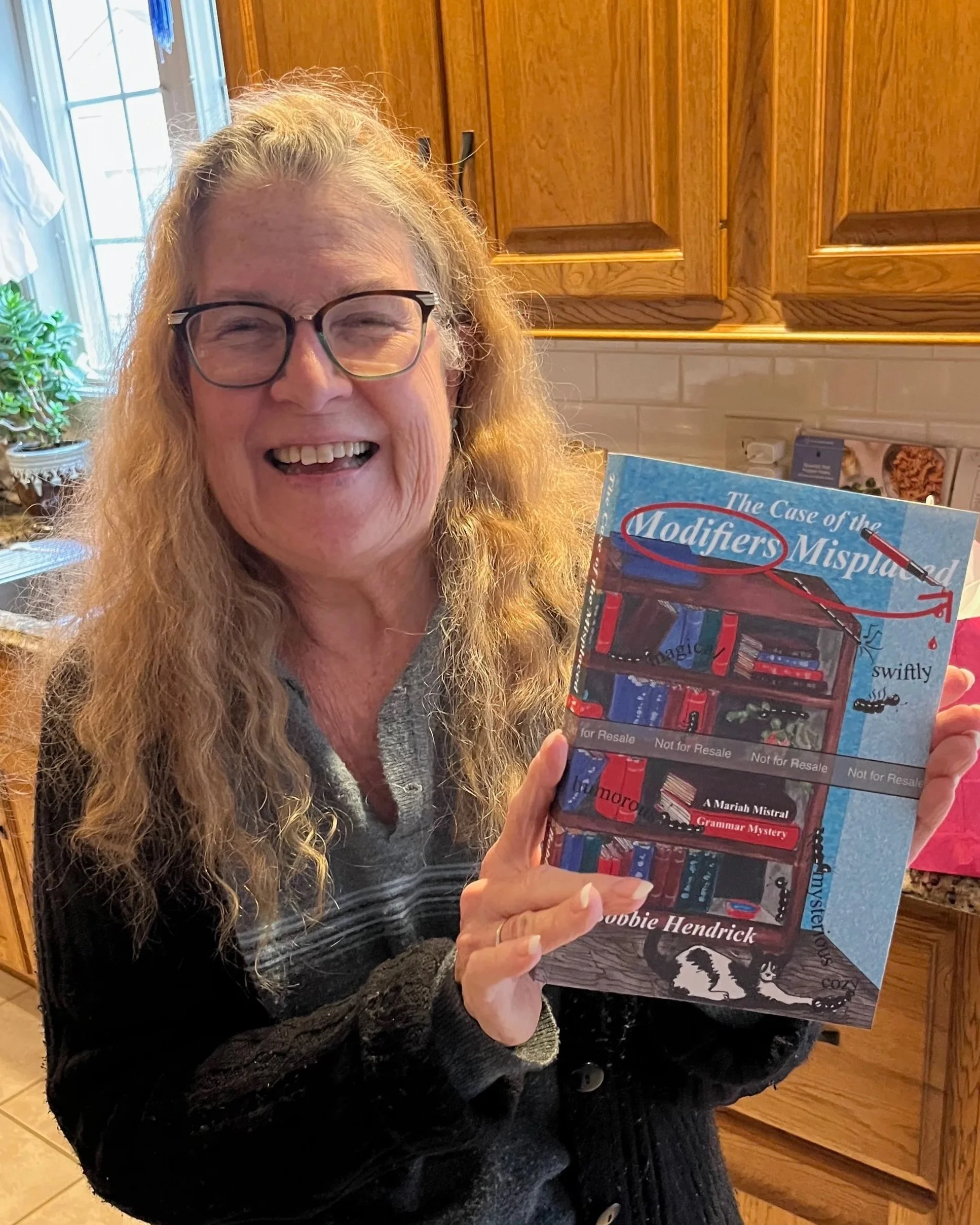 Woman with glasses smiling and holding a book titled 'The Case of the Modifiers Misplaced' in a kitchen with wooden cabinets and a window with plants.