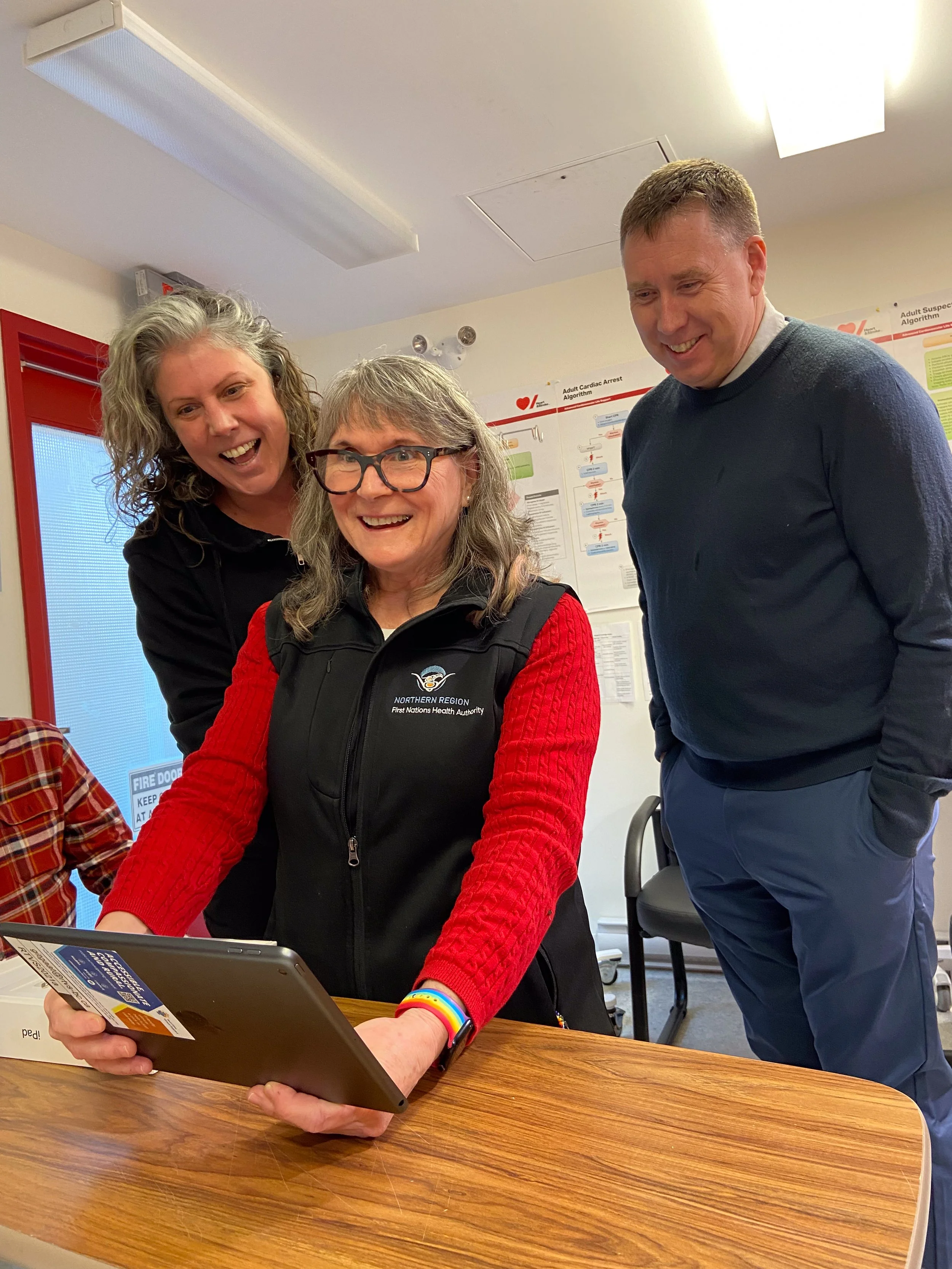 Three people smiling and looking at a tablet in a classroom or office setting.