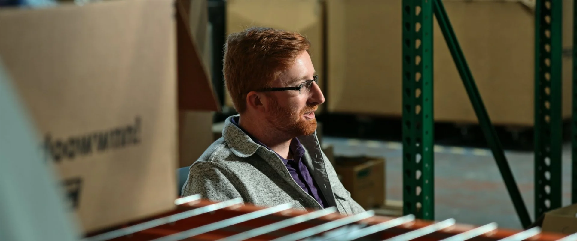 A man with red hair and glasses sitting in a warehouse surrounded by cardboard boxes and green metal shelving units.