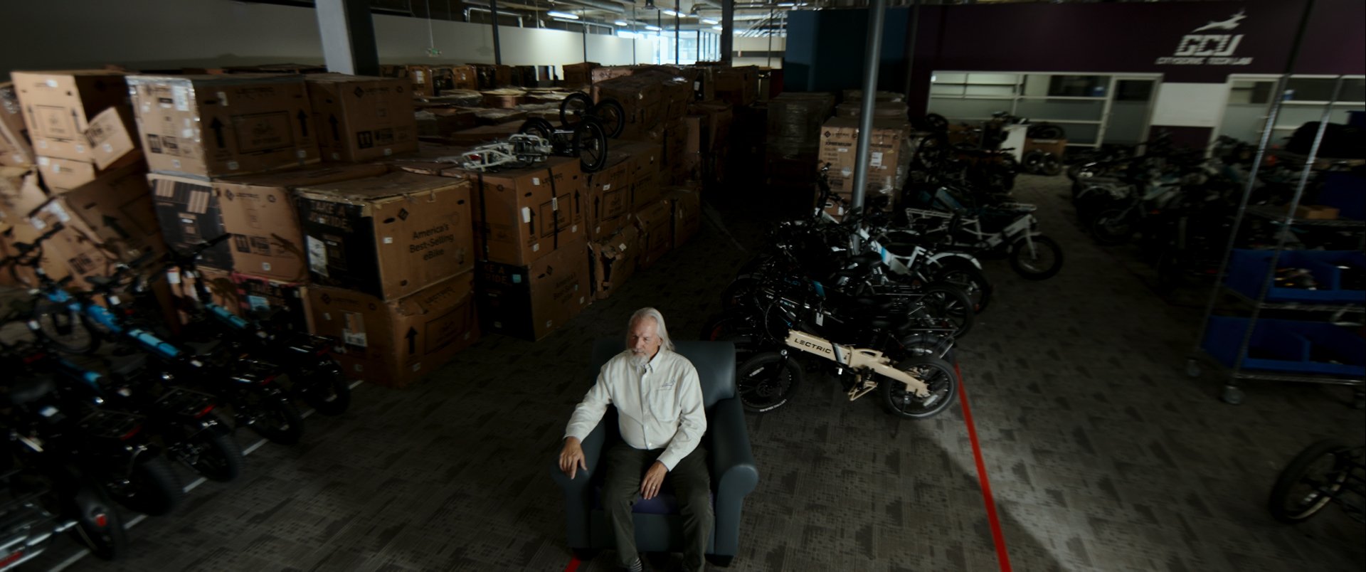 A man sitting in a dark room filled with stacked boxes and bicycles, with motorbikes and additional bikes on the right side.