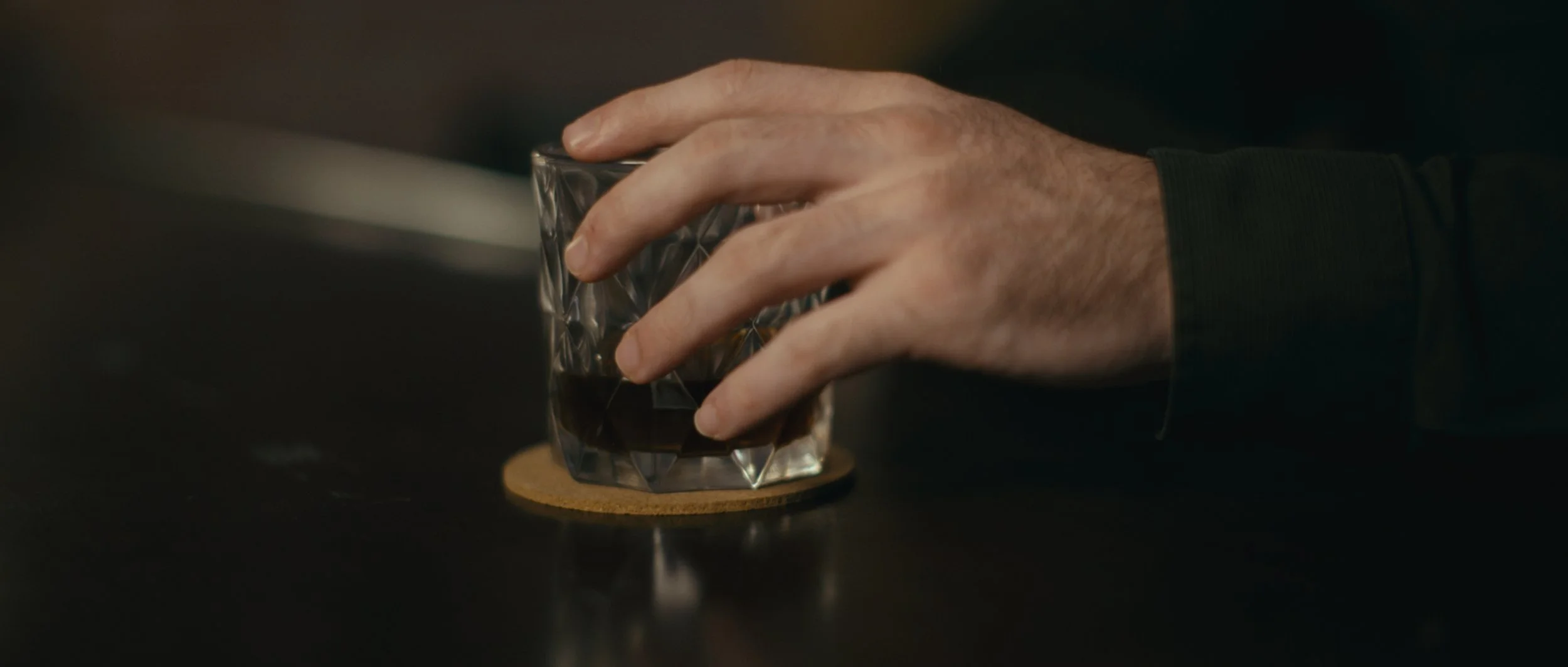 A person's hand holding a glass of dark liquid on a coaster, resting on a dark surface.