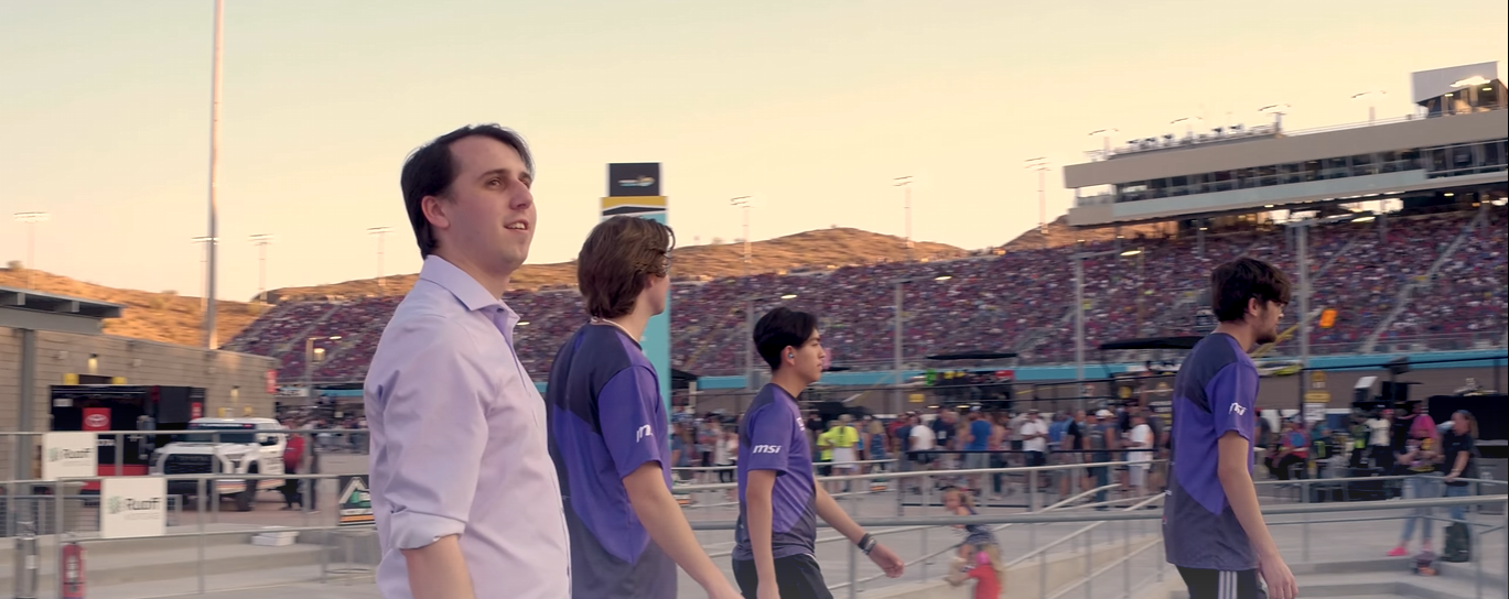 Four young men walking in a racing stadium, with a large crowd in the stands and various equipment and tents in the background.