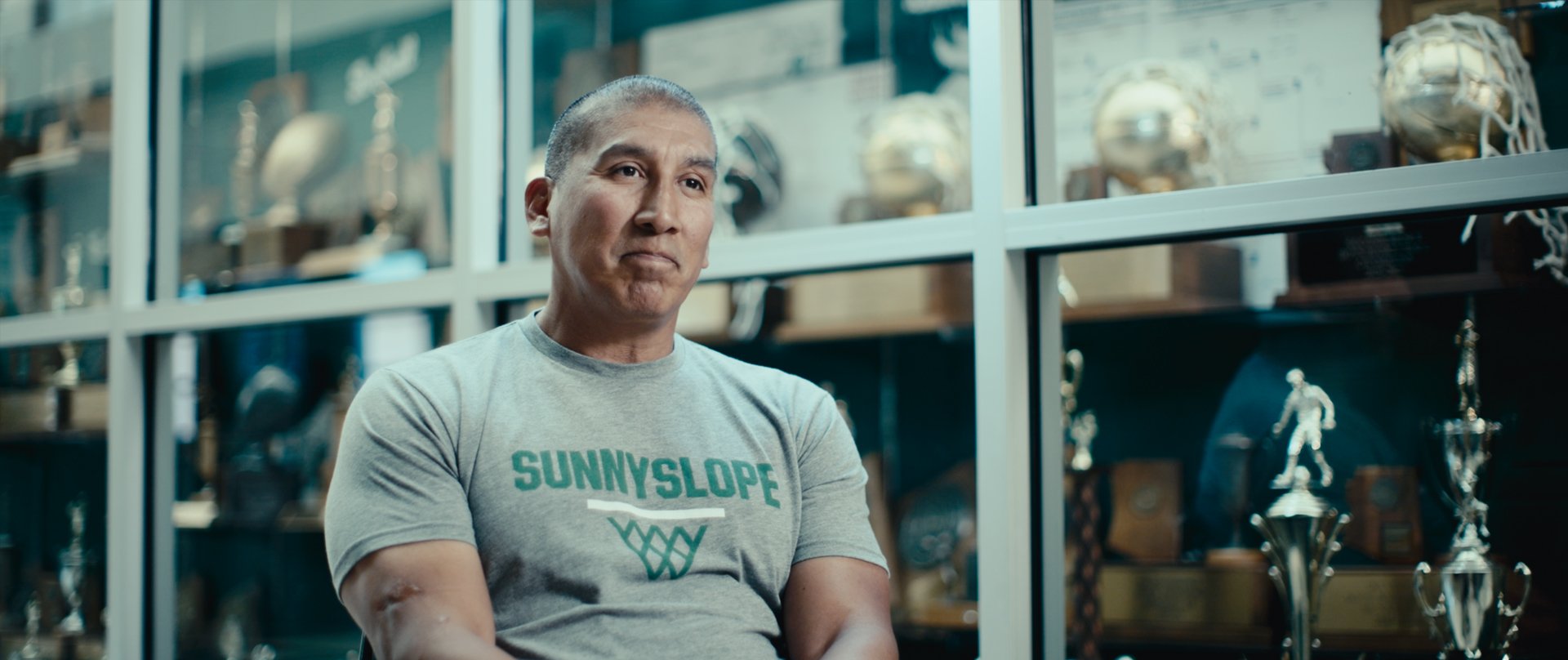 A man with a shaved head and light skin wearing a gray T-shirt with 'Sunny Slope' and a basketball hoop logo, sitting in front of a glass display case filled with trophies and awards.