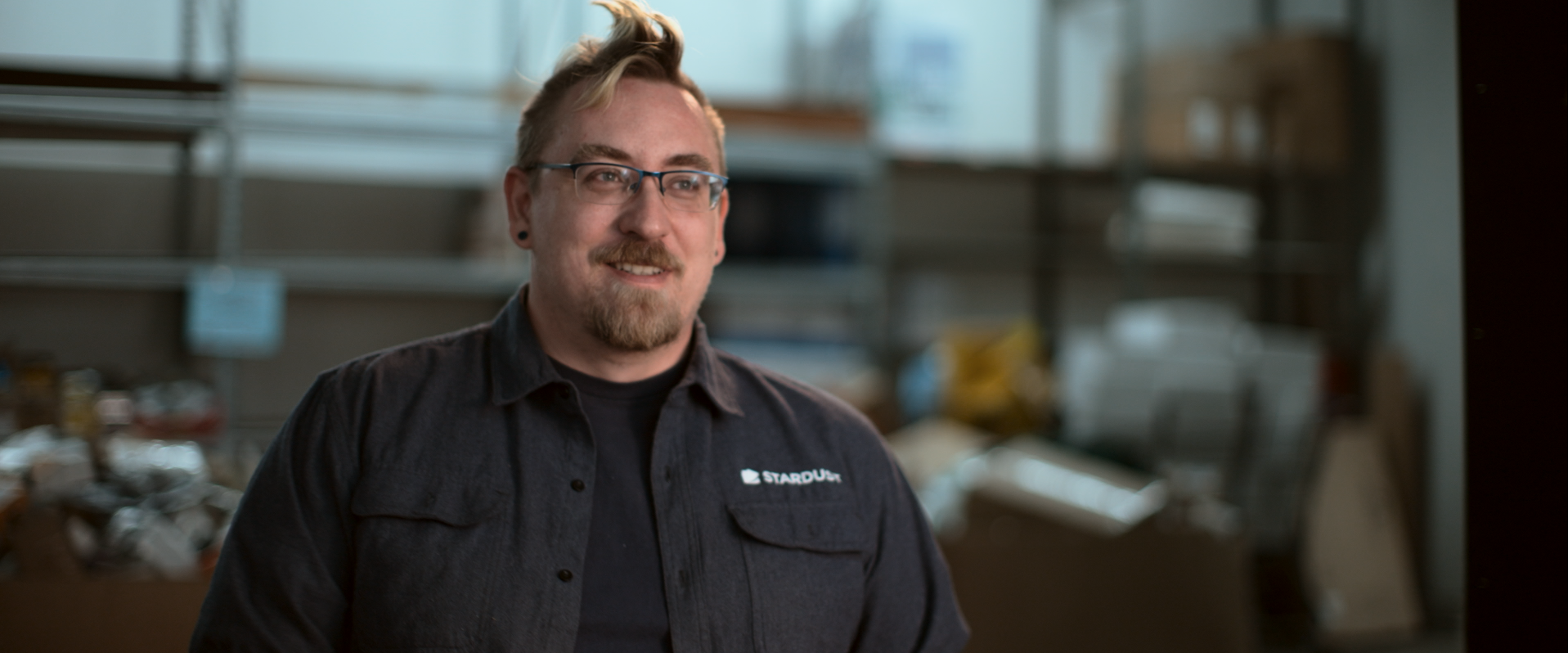A man with glasses, a beard, and a fashionable hairstyle smiling in an industrial or workshop setting.