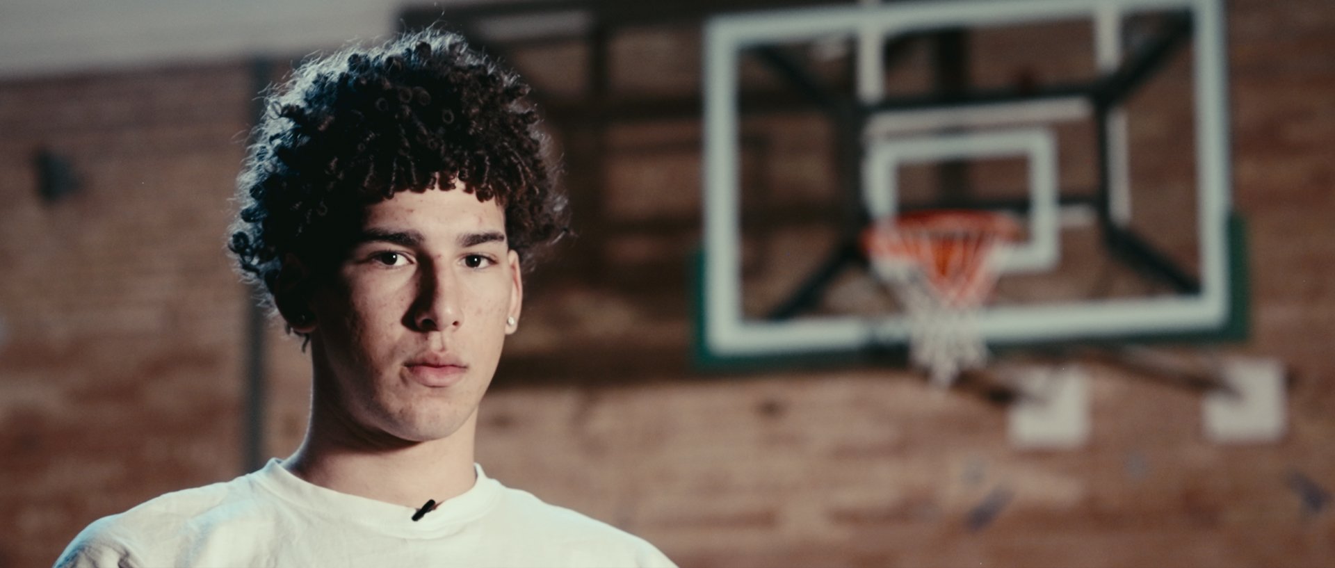 A young man with curly hair and earrings standing in front of a basketball hoop in a gym or sports court with a brick wall background.
