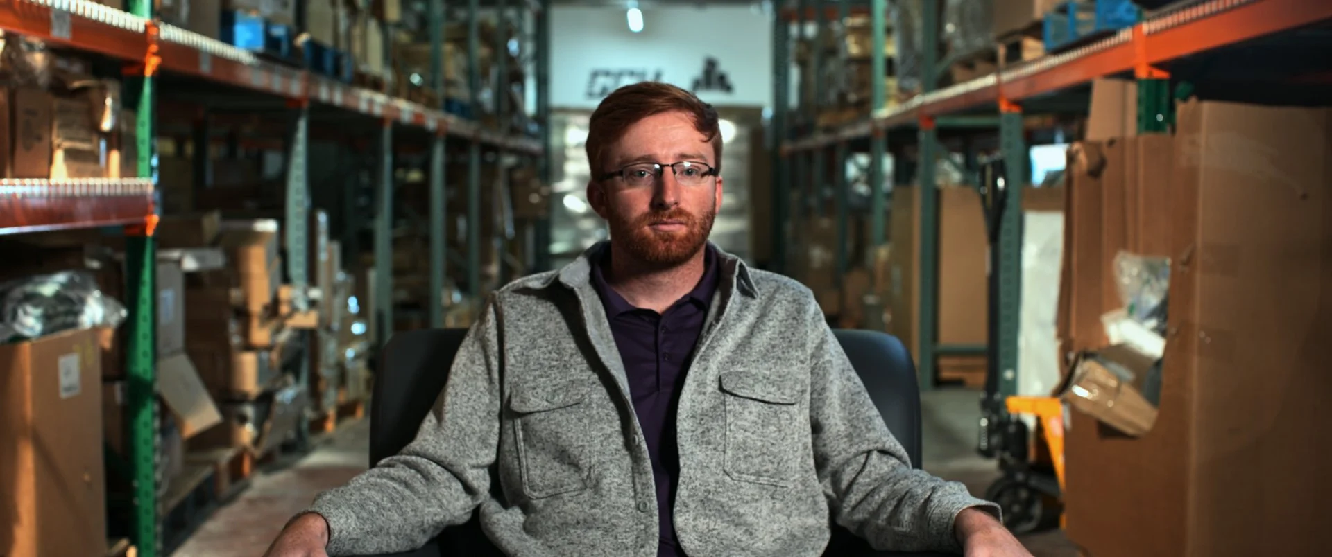 A man with glasses and a beard sitting in a warehouse surrounded by shelves filled with boxes and items.