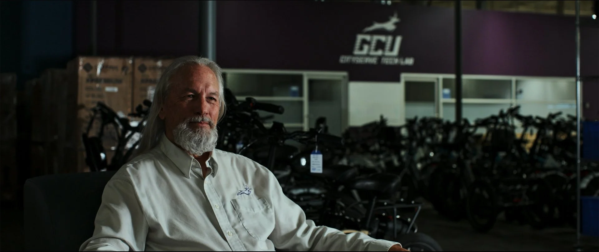 An older man with long gray hair and a beard, wearing a white button-up shirt, sitting in a dark room with bicycles and a sign with a deer logo in the background.