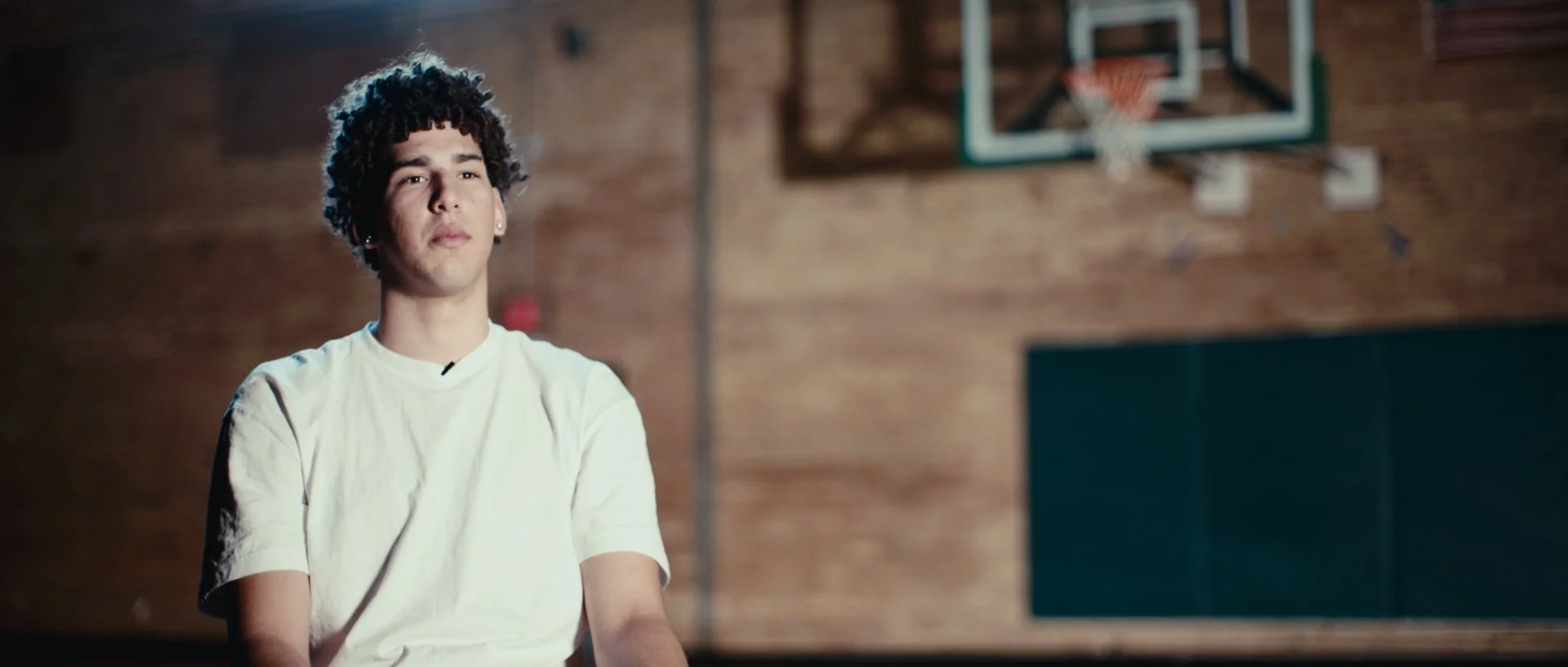 Young man with curly hair wearing a white t-shirt, sitting in a gymnasium with a basketball hoop and brick wall in the background.