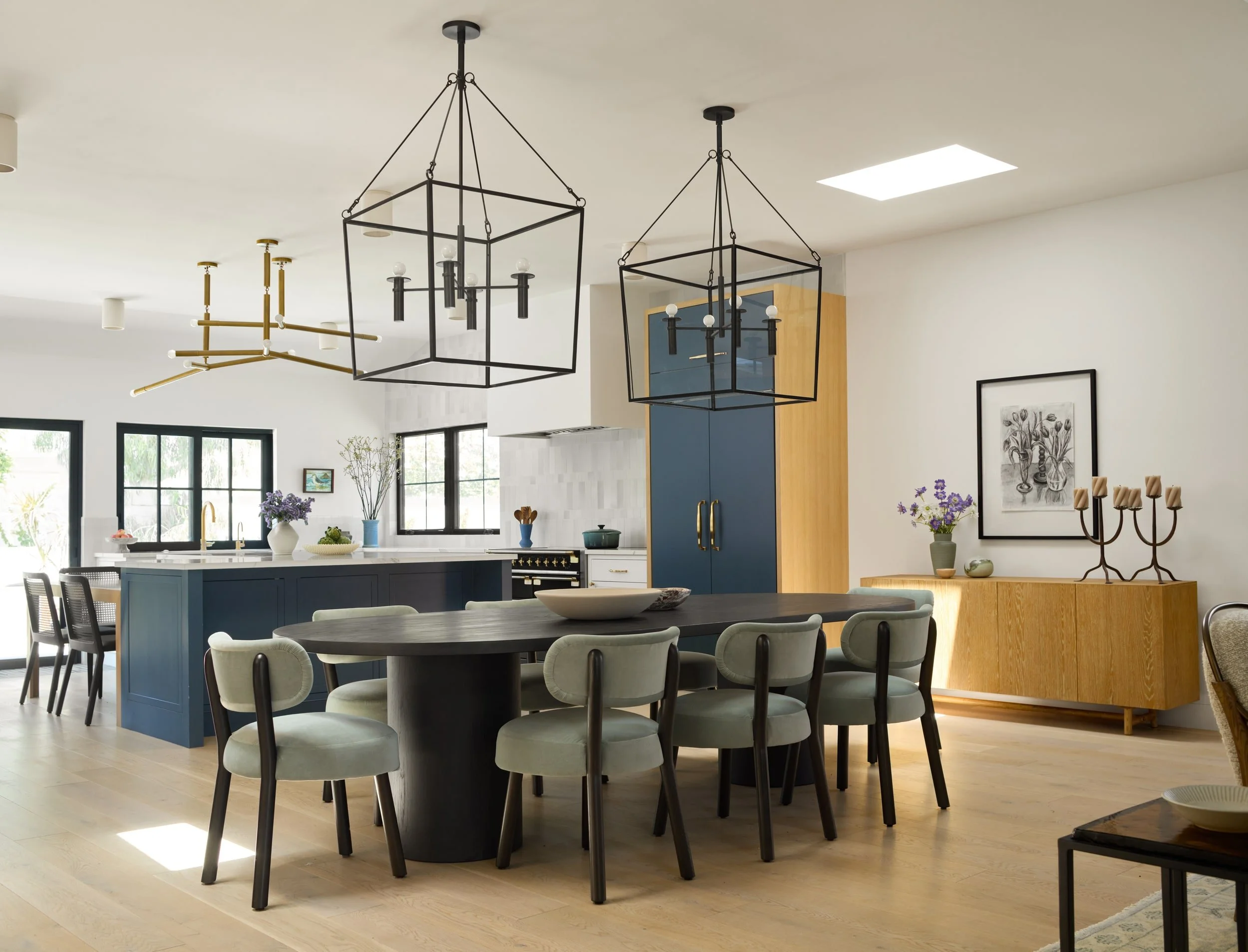 Dining room with black geometric lantern pendants, round dark wood table and curved sage chairs beside a kitchen