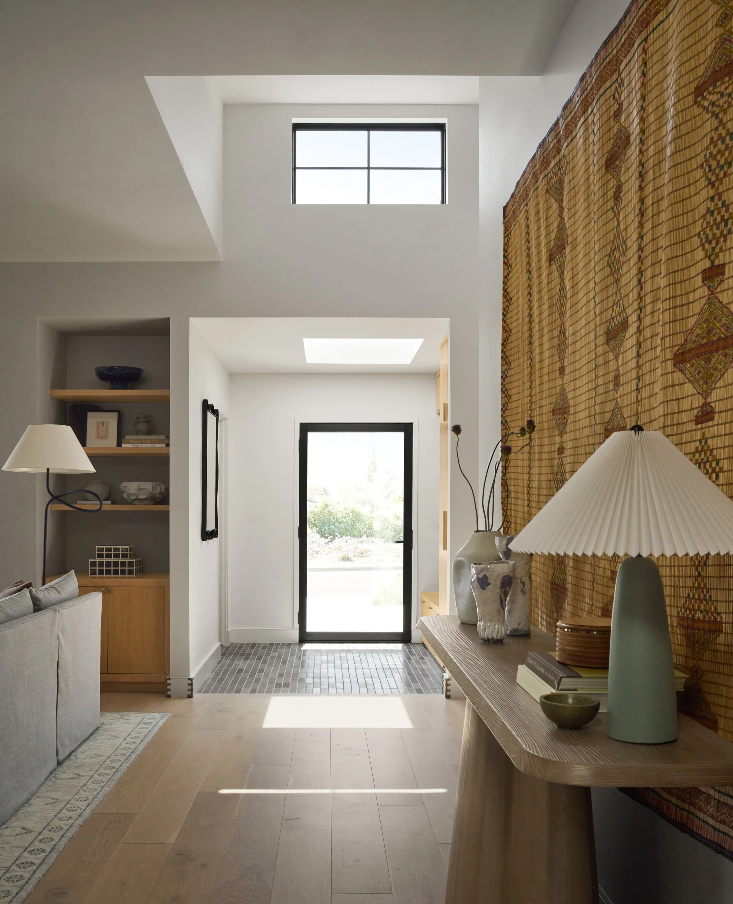 Entry hallway with a woven Tuareg mat wall hanging, sculptural wood console and pleated table lamp leading to the front door